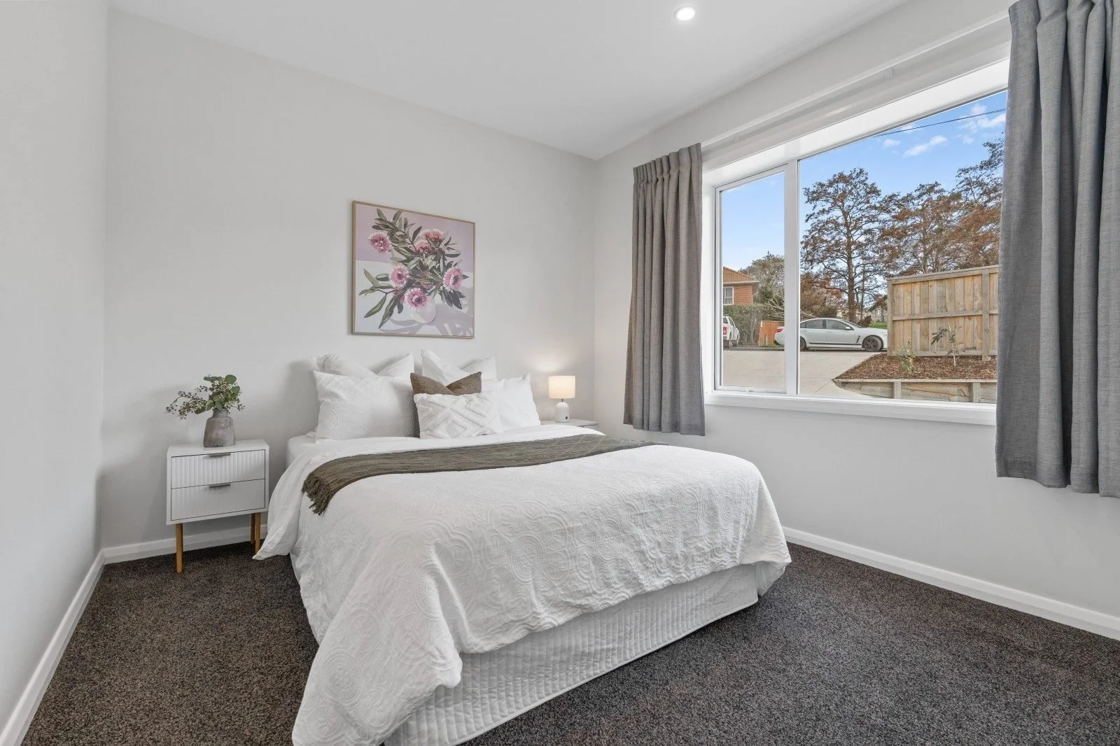 Interior of a bedroom with a white bed, gray curtains, a small nightstand with a plant, and a window showing an outside view with trees.