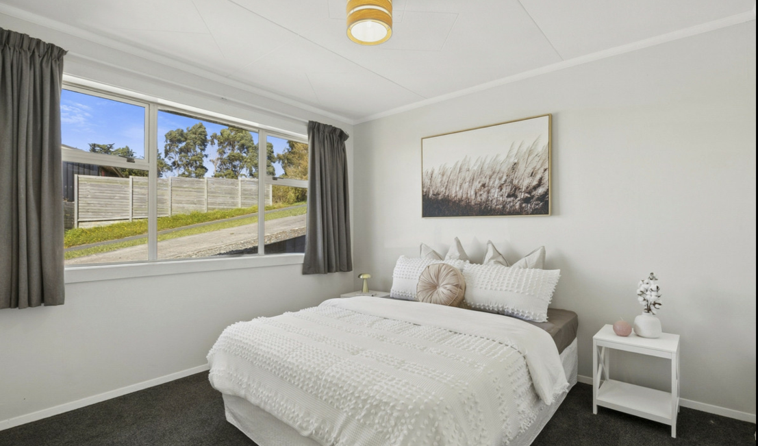Dunedin Home Staging - A cozy bedroom with a large bed dressed in white bedding, a round decorative pillow, and matching pillows. There is a small white bedside table with a cactus plant and a pink decorative object. Large windows with dark curtains 