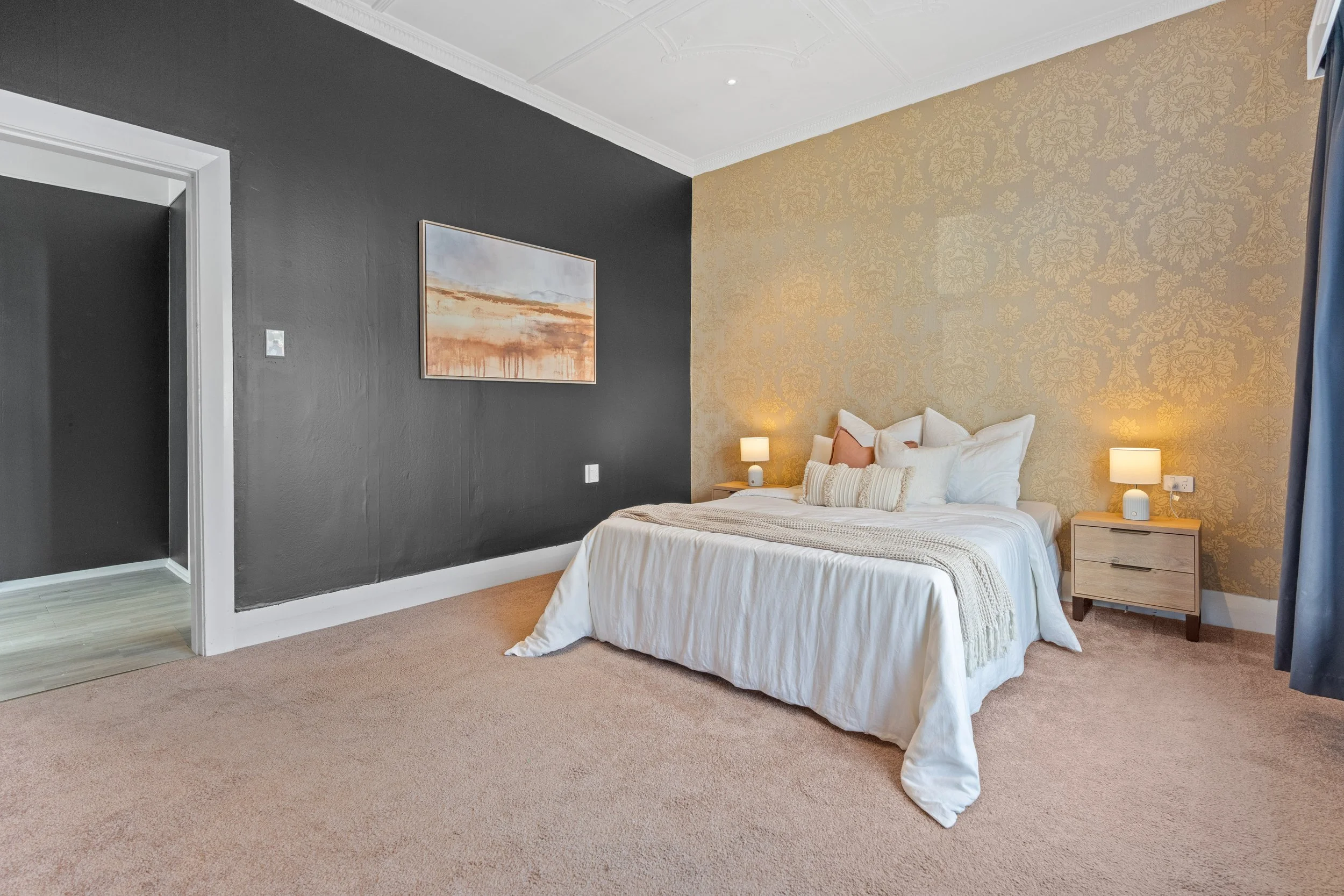 A bedroom with a bed, nightstand, and lamps, featuring a gold-patterned accent wall and a dark accent wall with a framed landscape painting.
