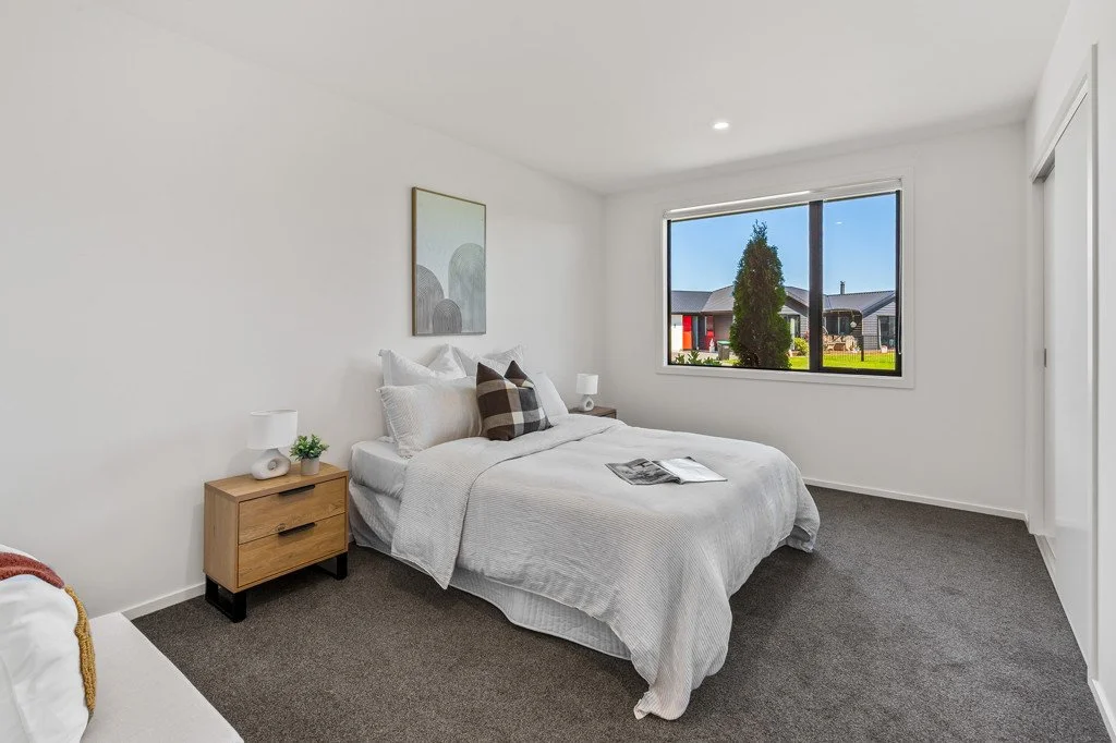 A modern bedroom with a bed, side tables, lamps, and a large window showing a suburban neighborhood.