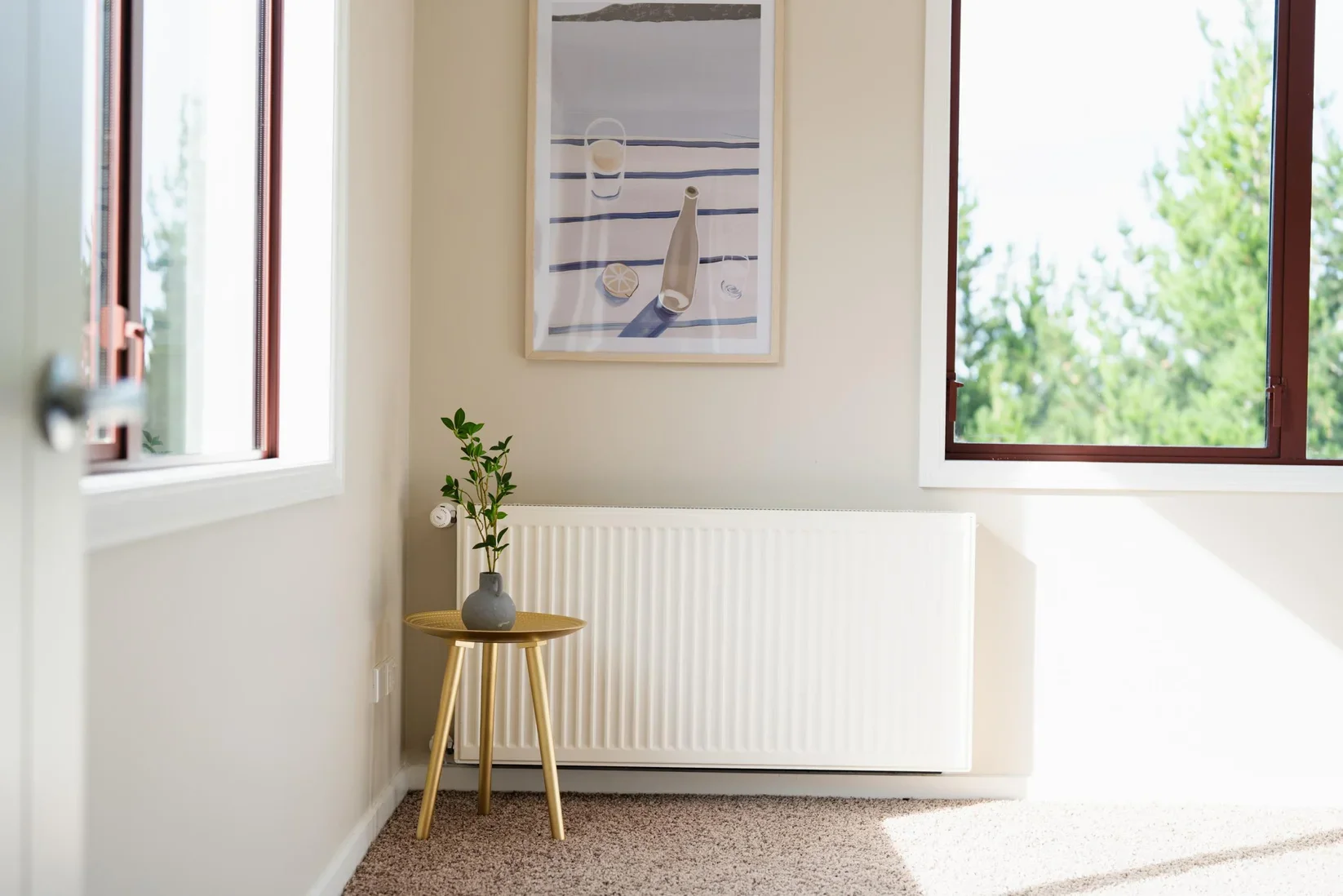 A corner of a room with beige walls and carpeted floor, featuring an open window showing green trees outside, a white radiator below a framed beach-themed art print, and a small round wooden side table with a gray vase holding a green plant.