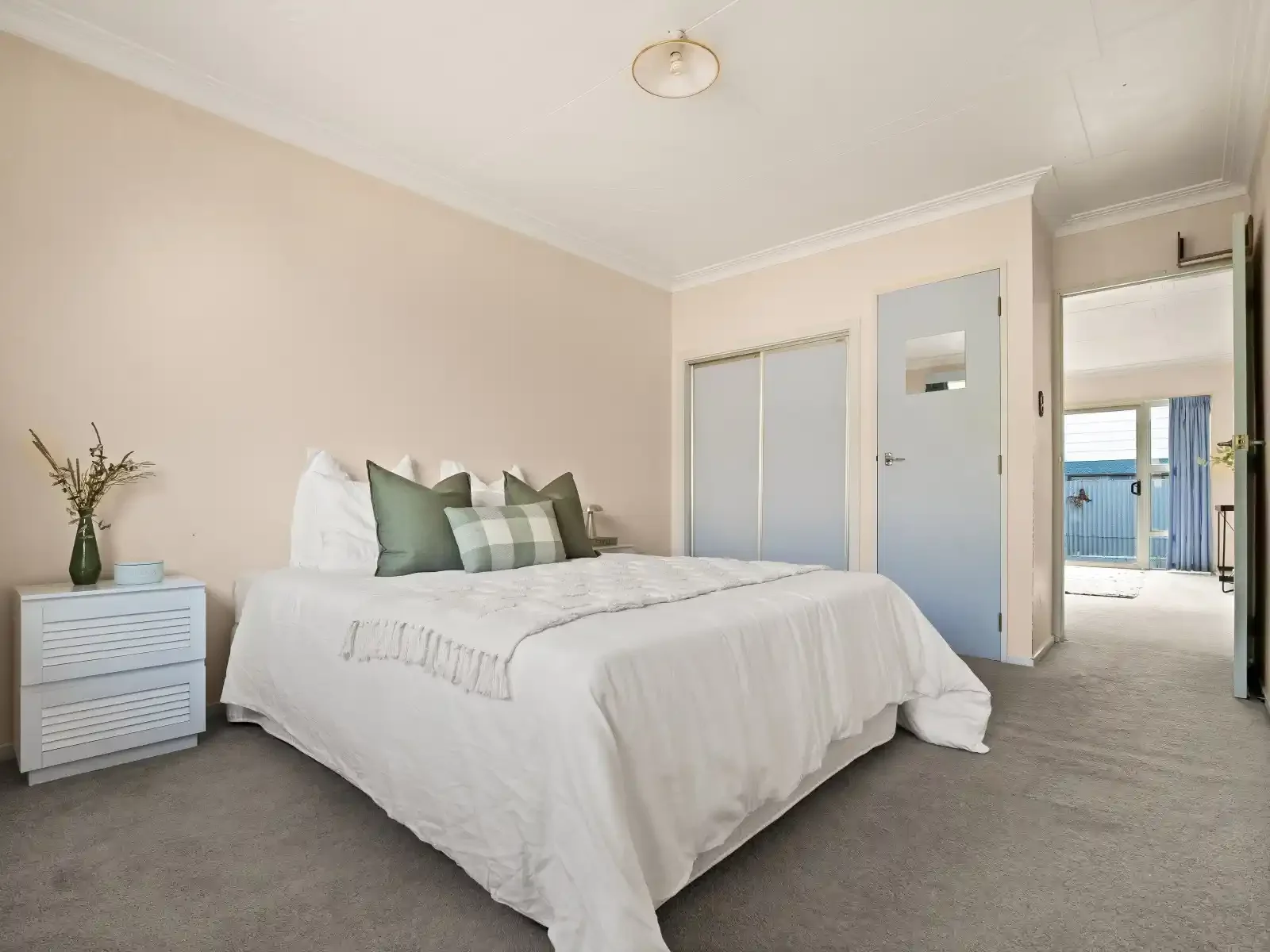 A bedroom with a large bed, white bedding, and green and patterned throw pillows. To the left of the bed is a white nightstand with a green vase with dried flowers. The room has beige walls.