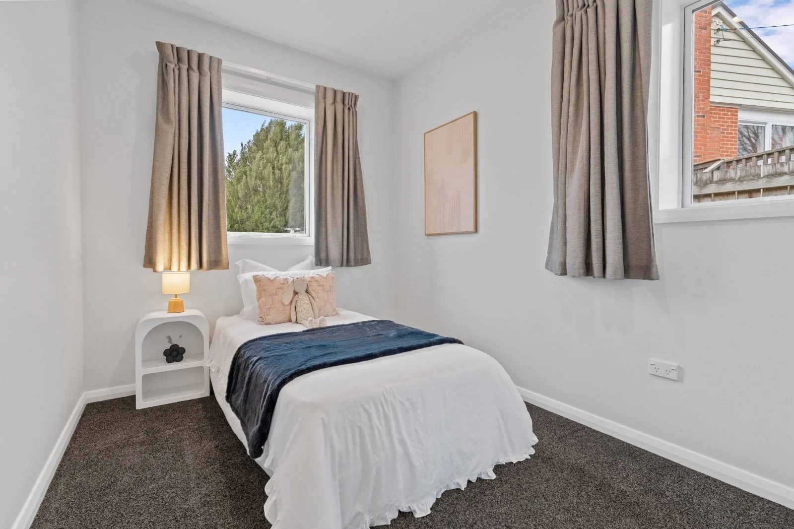 Small bedroom with a white bed, beige curtains, a nightstand with a lamp and a stuffed bunny, and two windows showing a brick house and trees outside.