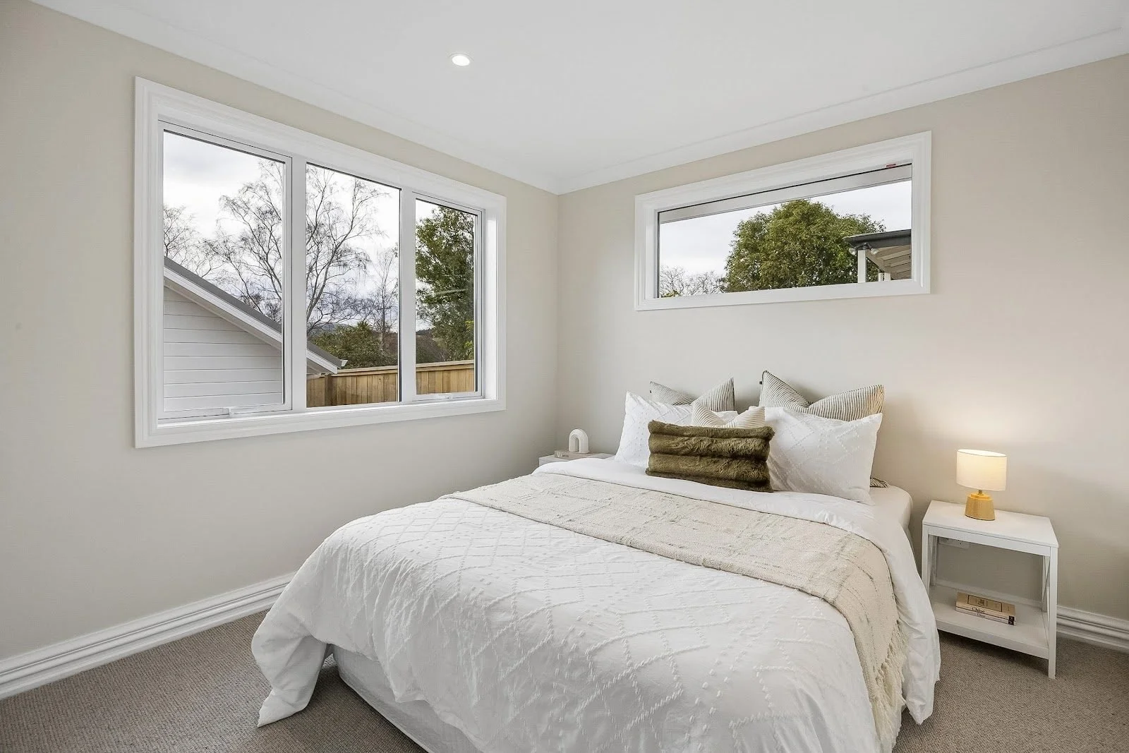 Bedroom with white bedding, pillows, nightstand with lamp, and large windows showing trees outside.