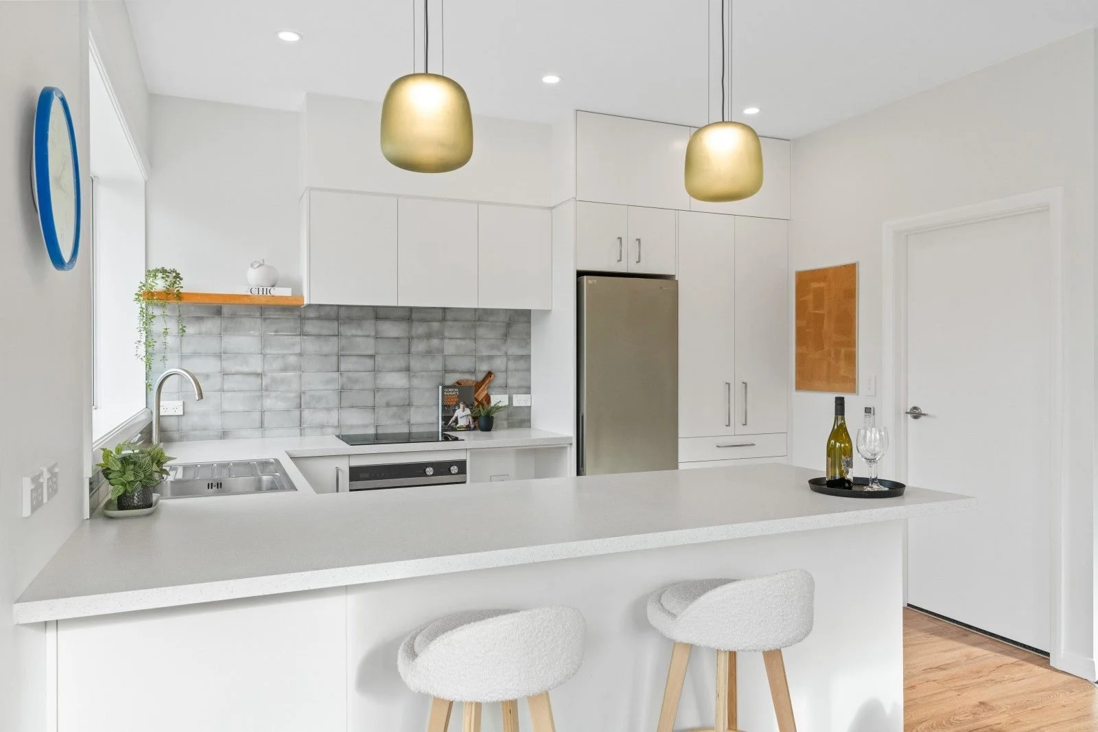 Modern kitchen with white cabinets, gray backsplash, stainless steel refrigerator, and a white countertop island with two light-colored bar stools. Decorative items include a wine bottle and glasses, a small plant, and a hanging pendant light.