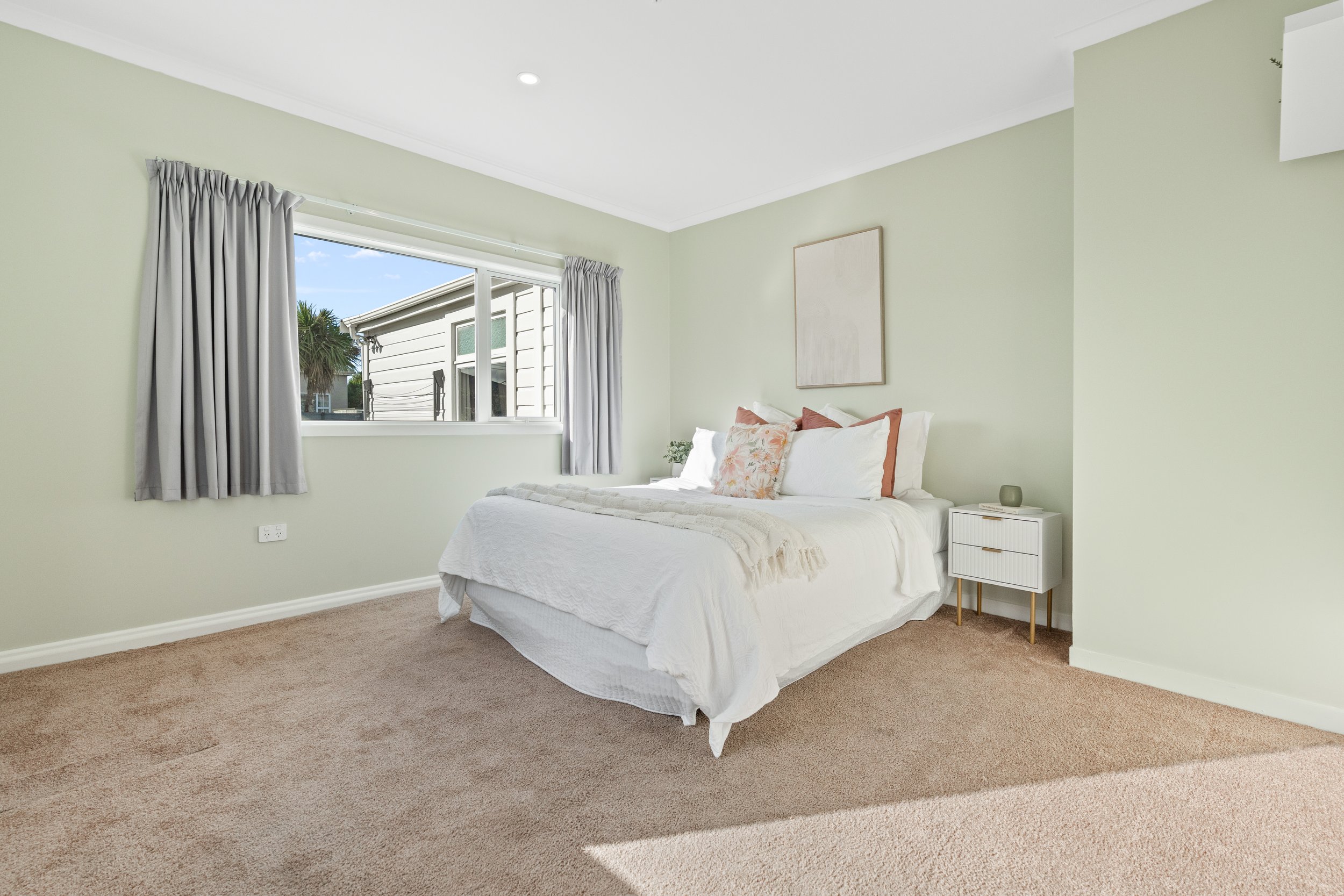 A neatly made bed with white bedding and decorative pillows in a bedroom with light green walls, a window with curtains, a small bed-side table, and a framed art piece on the wall.