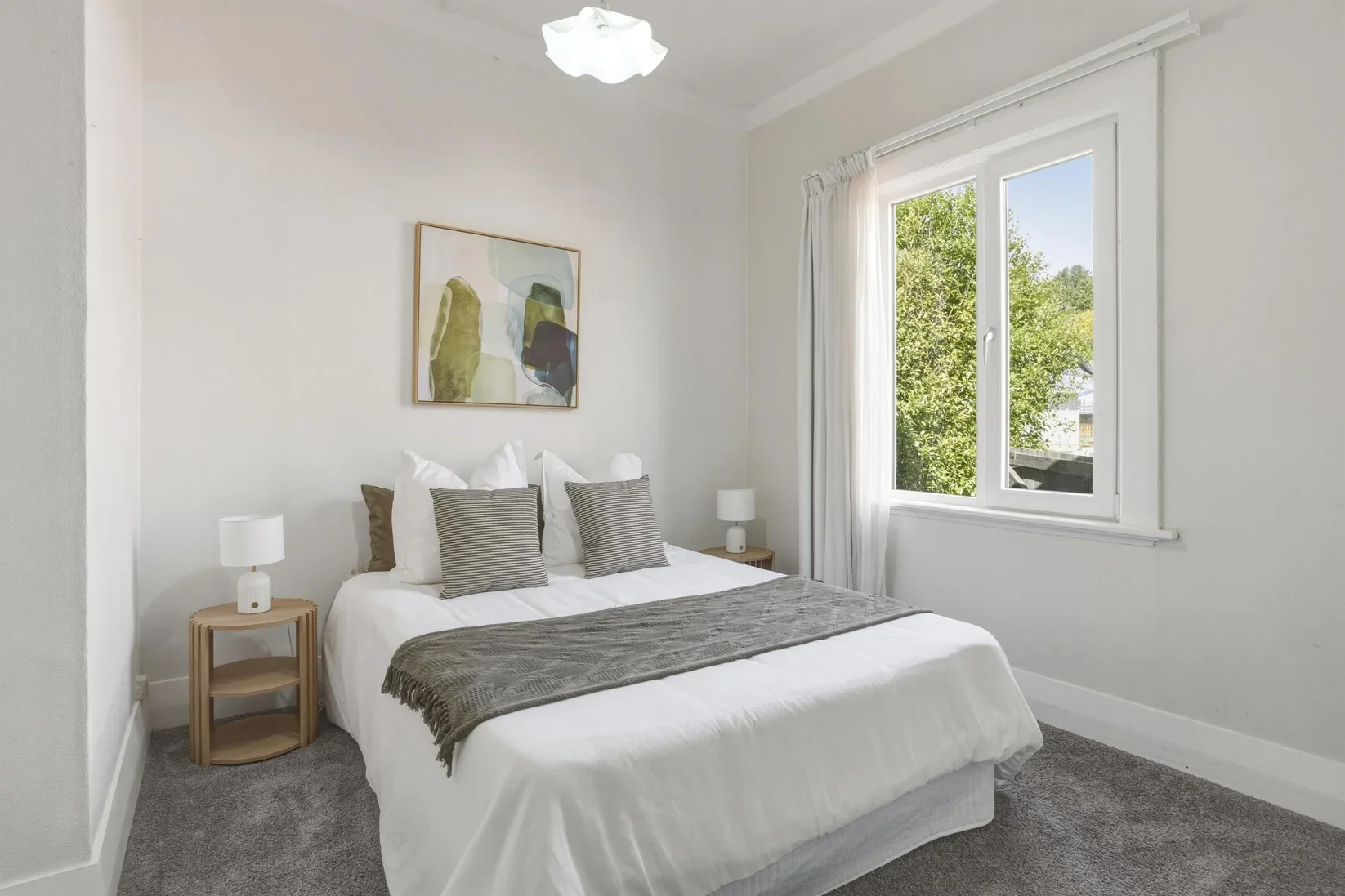 Home Staging Dunedin - A bright bedroom with a bed, two bedside tables with lamps, a large window with curtains, a piece of abstract art, and a ceiling light fixture.