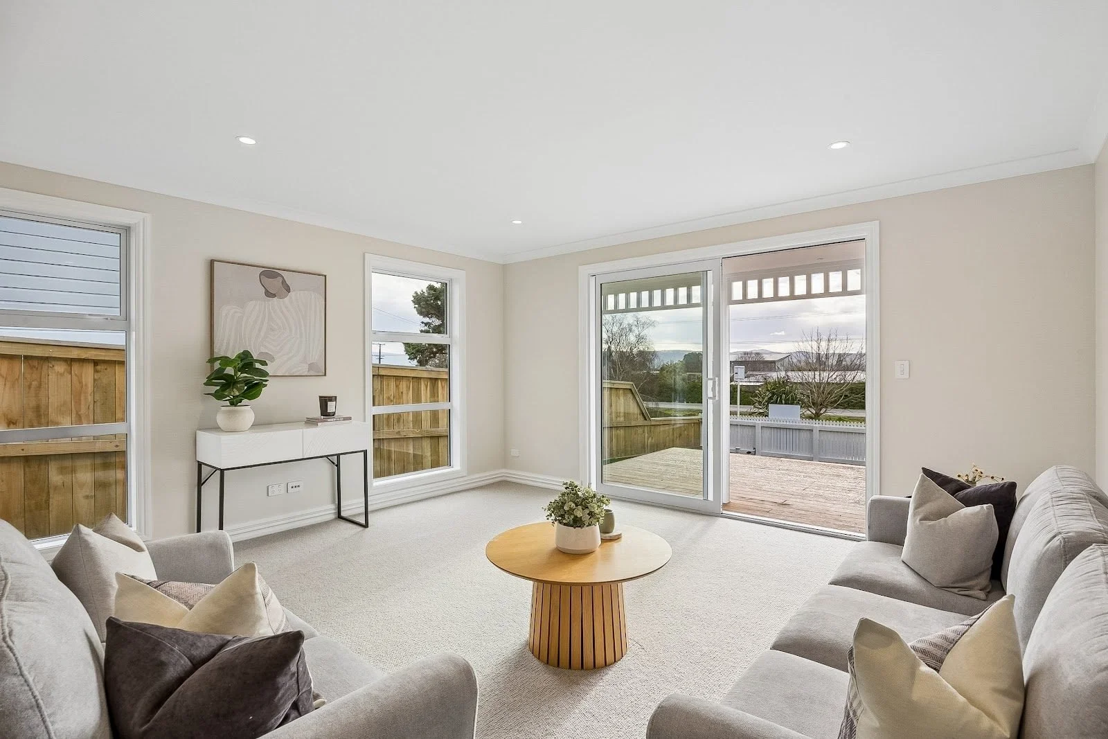 Living room with beige sofas, a wooden coffee table with a plant, large windows, and a sliding glass door leading to a deck with views of trees and a cloudy sky.