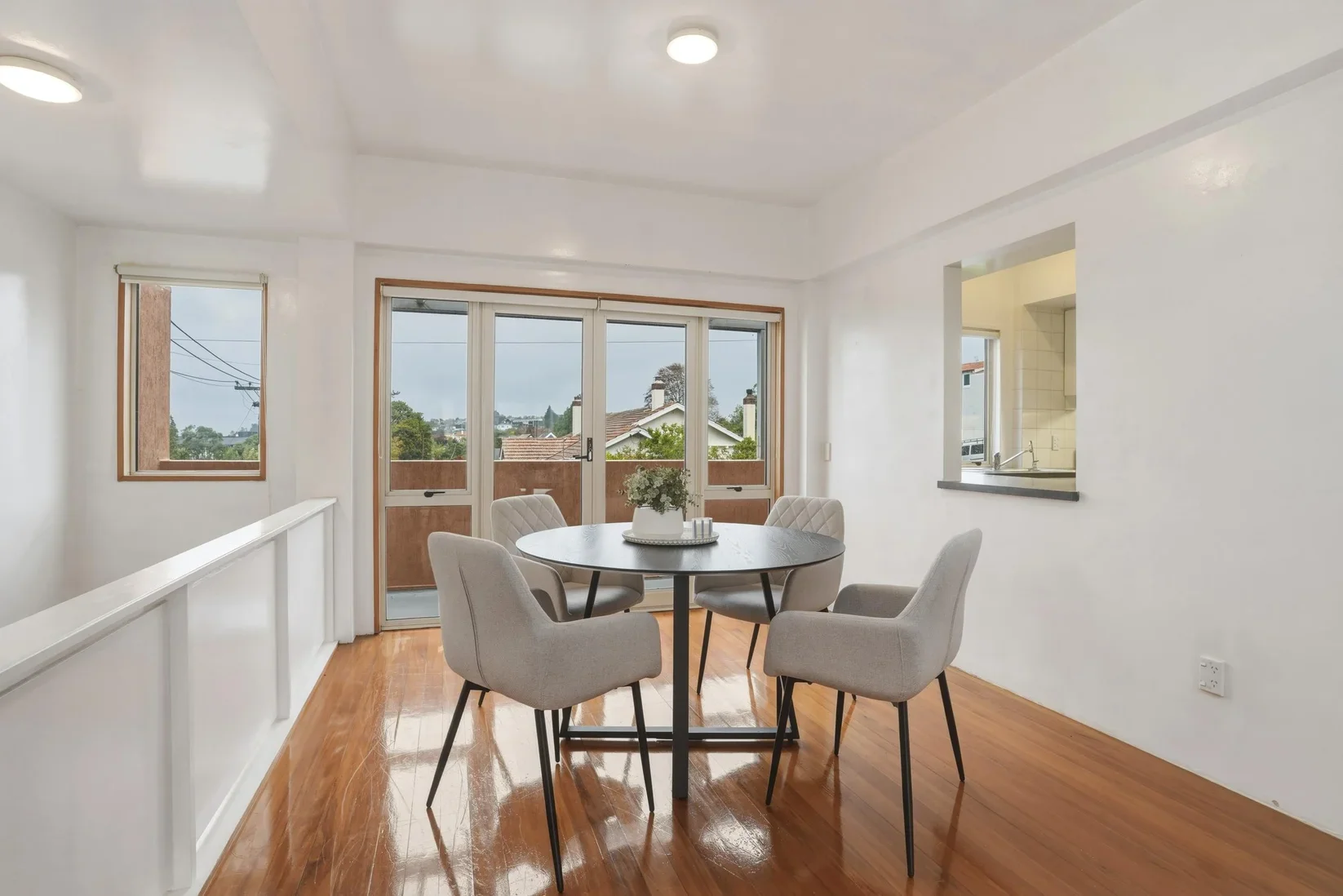 Home Staging Dunedin - Dining area with a round table and five upholstered chairs, large window and glass door leading to a balcony with a view of neighboring houses and trees, hardwood floors, white walls, and a partial view of a kitchen with a coun
