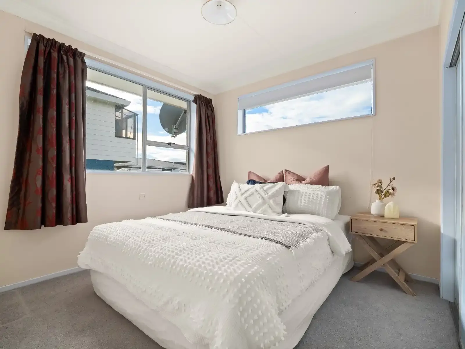 A bedroom with a large bed covered in white textured bedding and multiple pillows, a wooden nightstand with decorative items, a window with brown curtains, and a smaller horizontal window, with neutral-colored walls and carpeted floor.