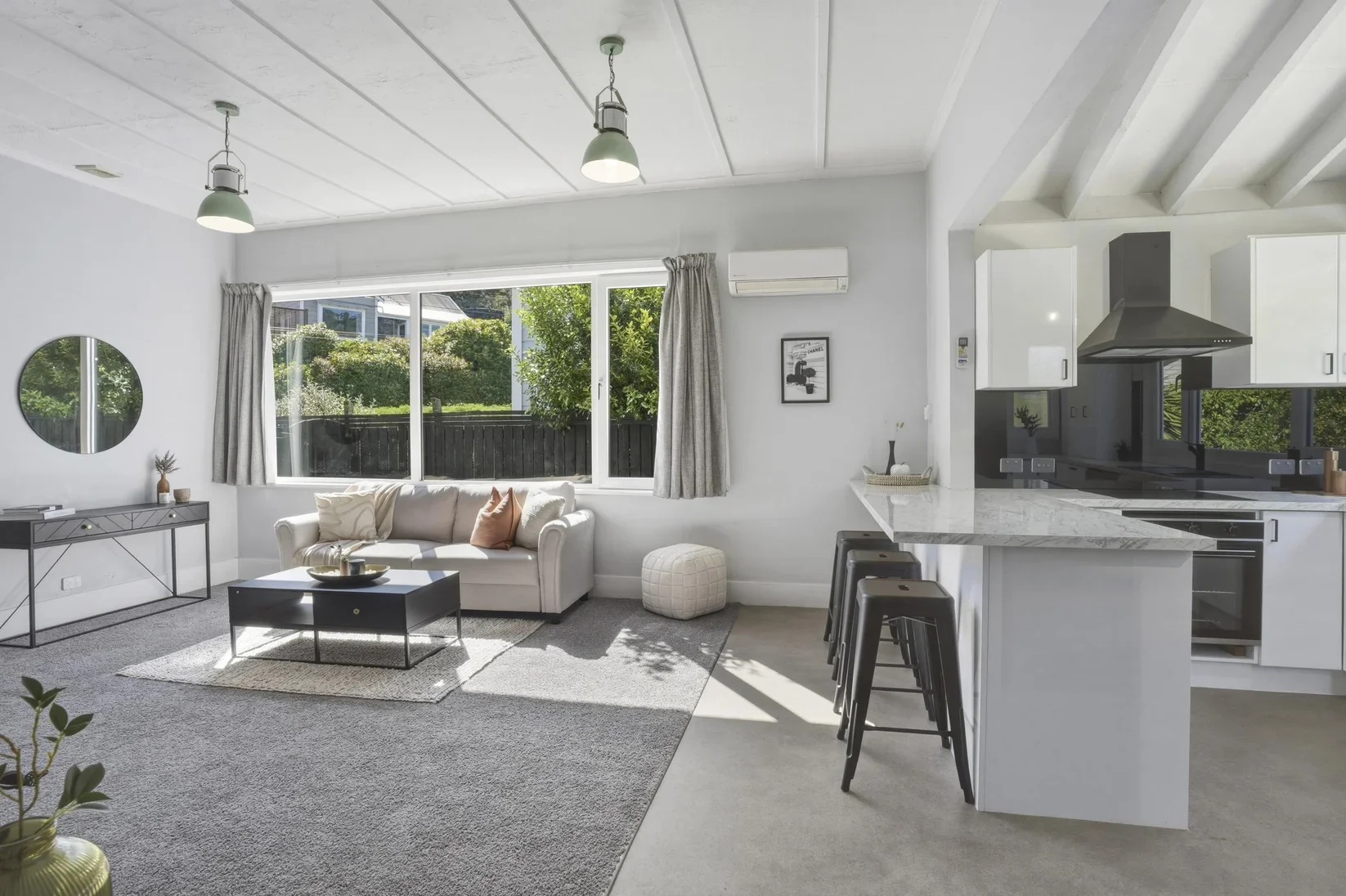 Bright living room with large window, white sofa with pillows, coffee table, decorative items, and a staircase with black bar stools near a kitchen with white cabinets and granite countertops.