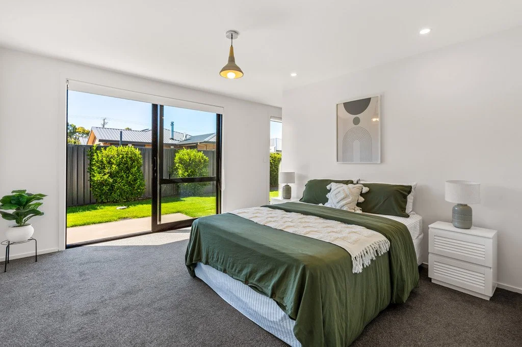 Modern bedroom with white walls, gray carpet, and large sliding glass door leading to a green backyard. The bed has white and green bedding, with two nightstands with lamps on either side. There is a large abstract artwork above the bed and a potted plant on the left.