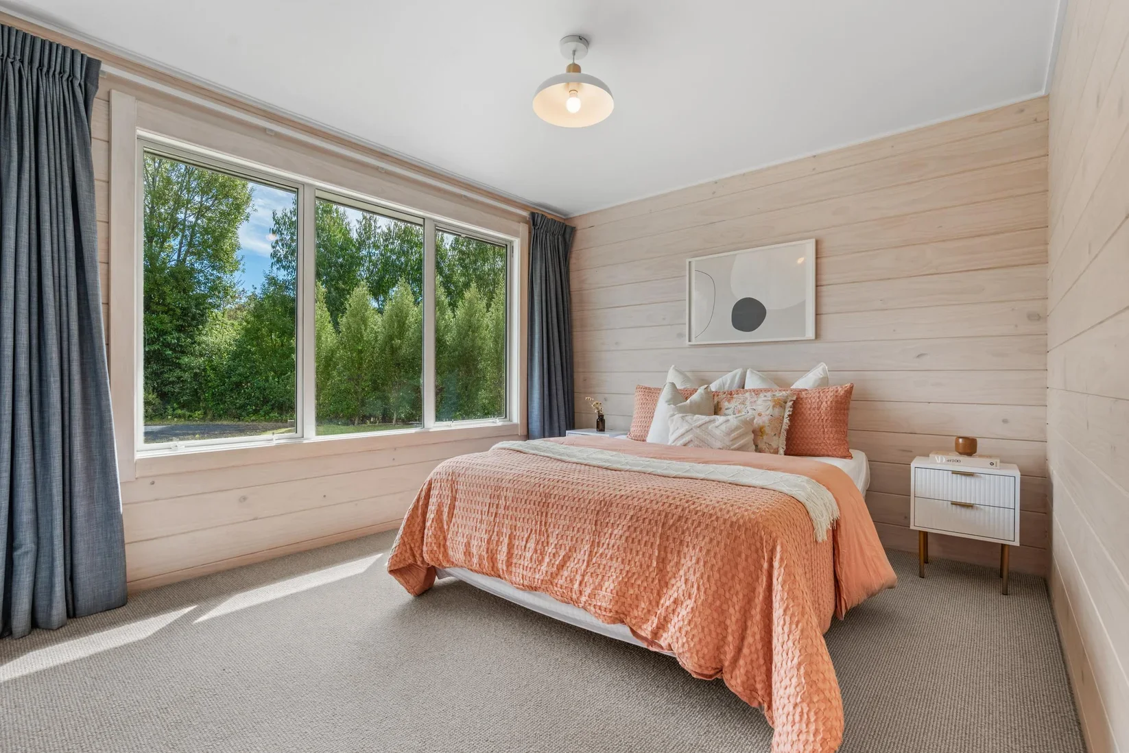 Property Staging Dunedin - A bedroom with a large window showing green trees outside, a bed with coral-colored bedding, white and patterned pillows, and a minimalist white nightstand with books and decor.