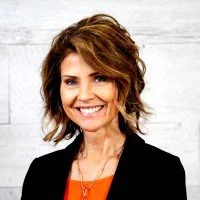 A smiling woman with shoulder-length, wavy brown hair, wearing a black blazer and orange top, standing in front of a white wooden background.