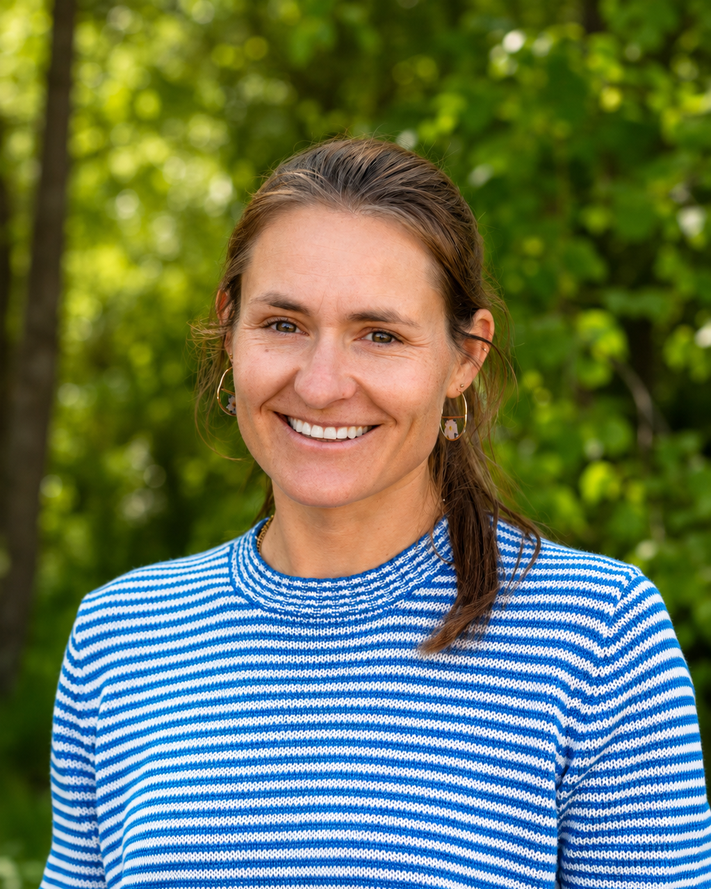 A woman smiling outdoors with a background of green trees, wearing a blue and white striped shirt and earrings.