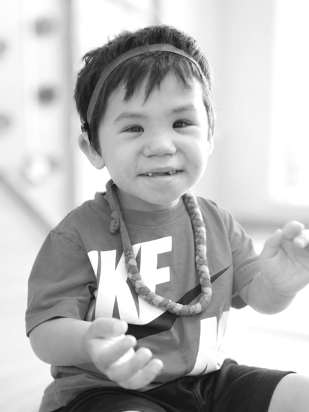 Black and white photo of a smiling young boy with short hair, wearing a headband, a T-shirt with large letters, a beaded necklace, and shorts.