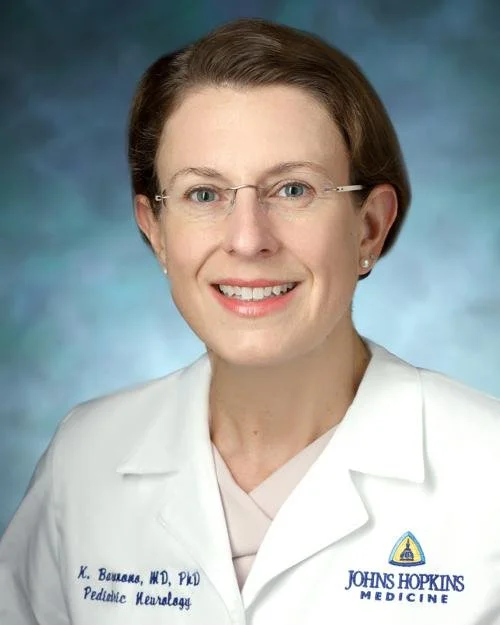 A woman with short brown hair, wearing glasses, smiling, dressed in a white medical coat with Johns Hopkins Medicine logo, and a name badge that reads 'K. Vahaba, MD, PHD, Pediatric Neurology'