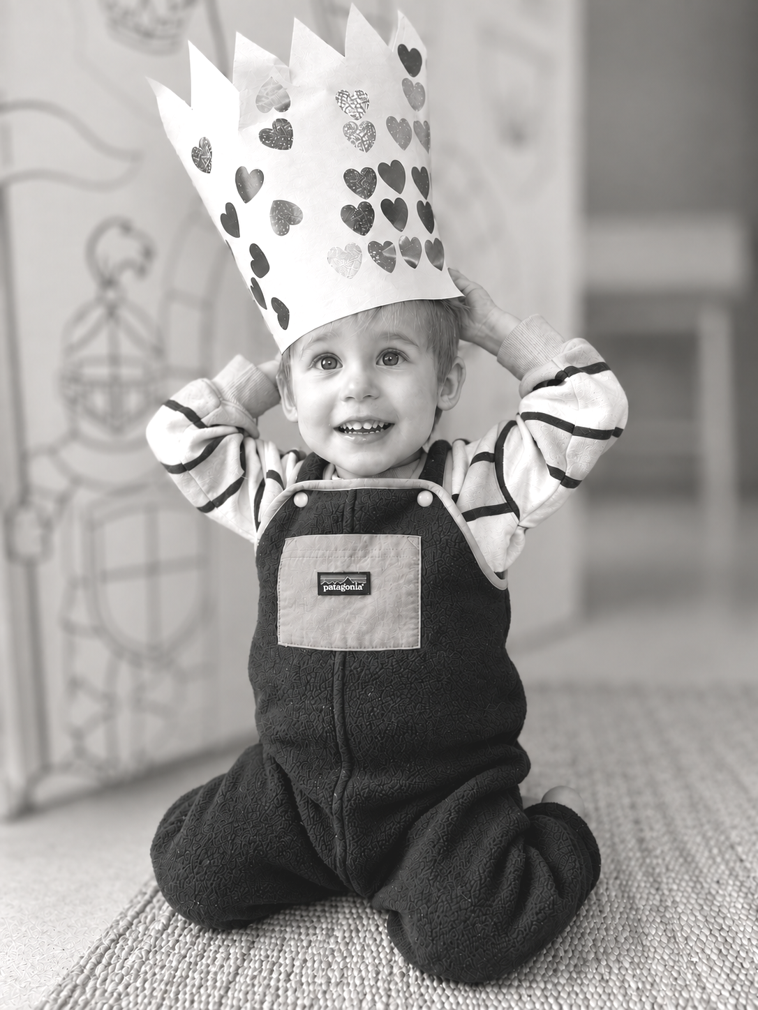 A young boy sitting on a textured mat, wearing a black overalls with a Patagonia logo, striped long-sleeve shirt, and a paper crown decorated with heart shapes, smiling and holding the crown on his head.