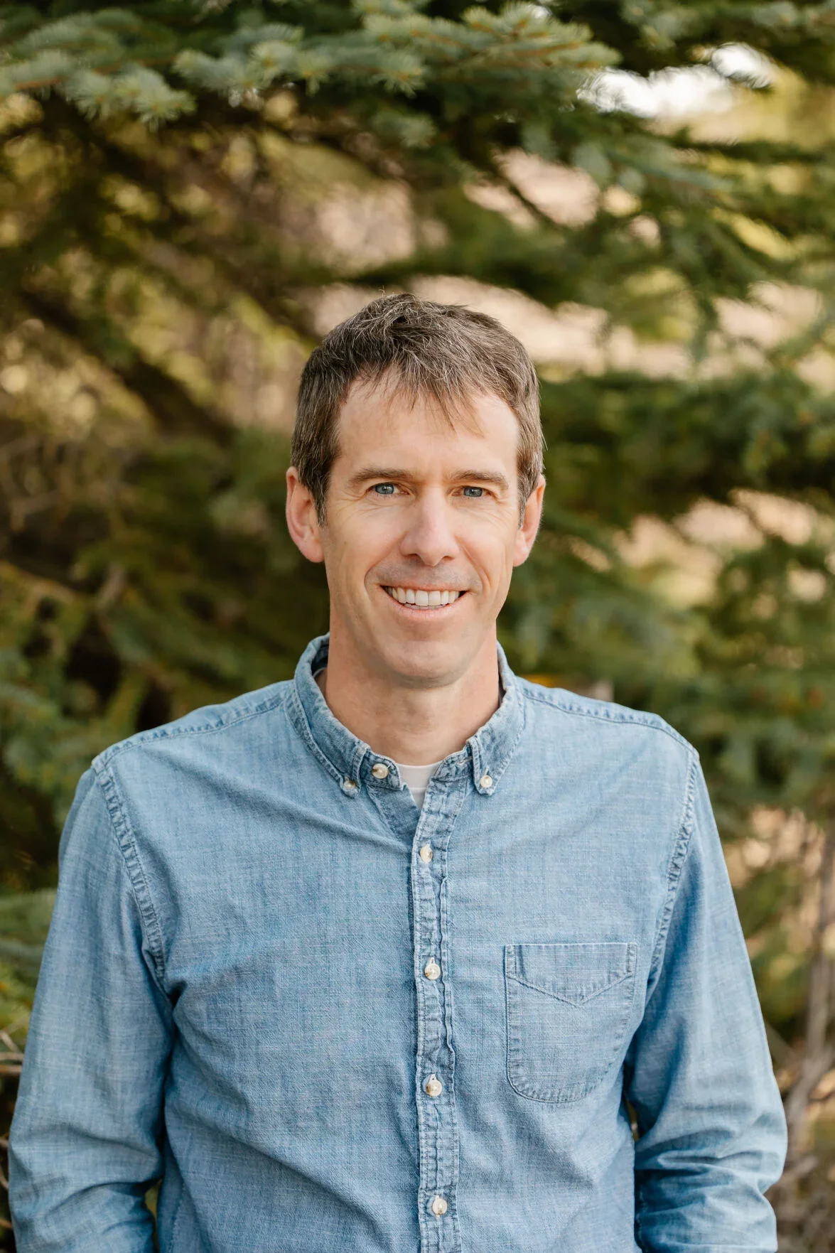 A smiling man with short brown hair wearing a light blue button-down shirt, standing outdoors in front of green trees.
