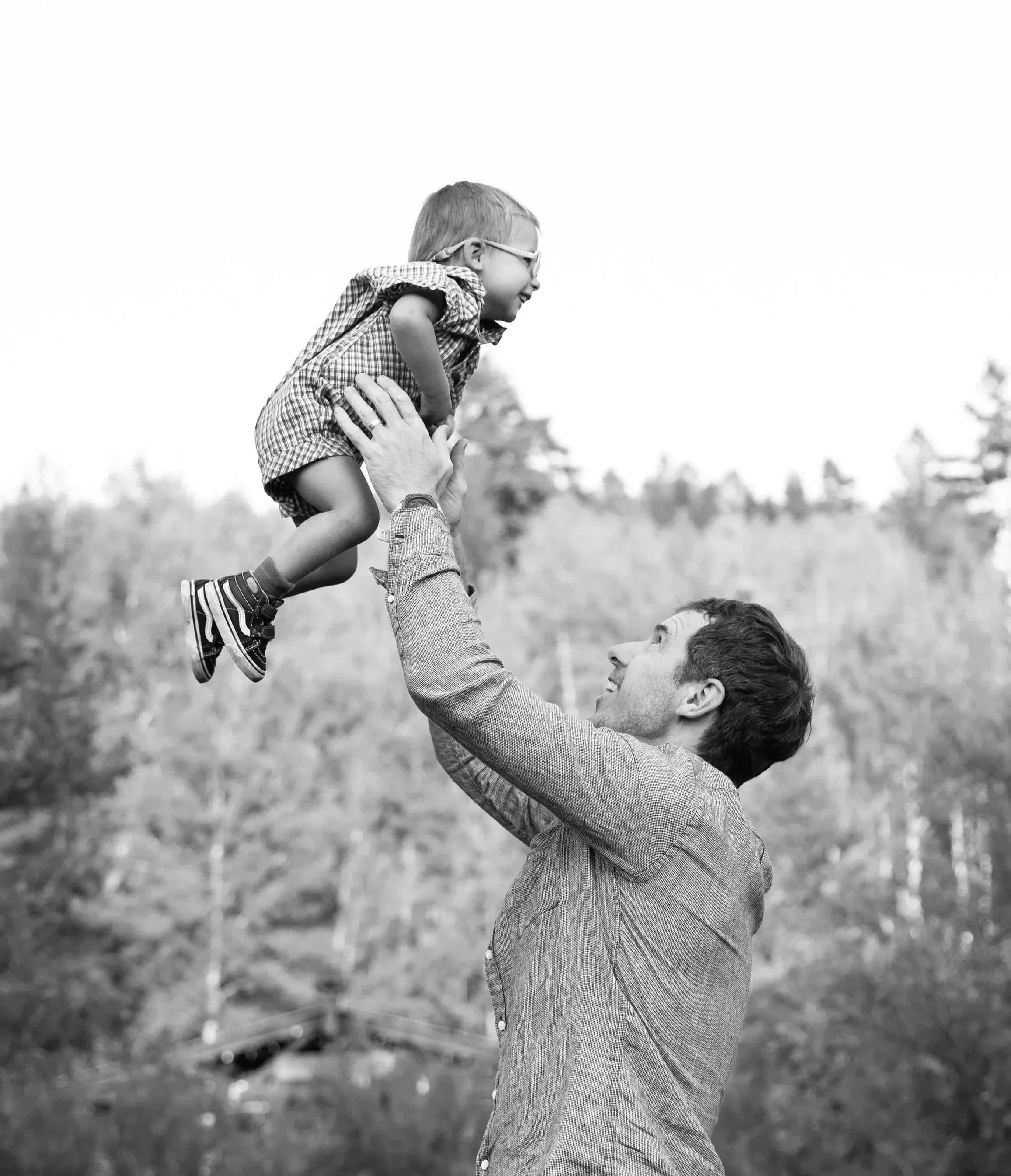 A man lifts a young boy in the air outdoors, both smiling. The boy wears glasses and a checkered shirt, and the background is blurry, indicating a natural setting.