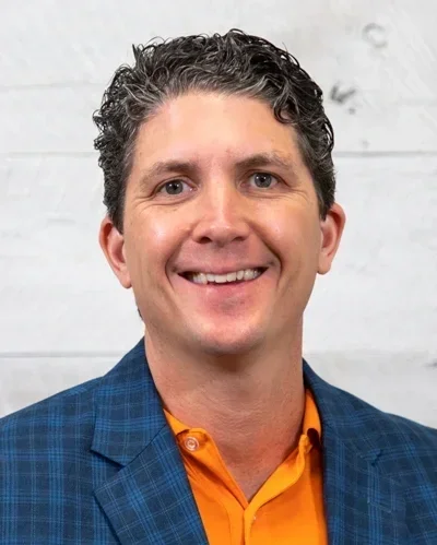 A smiling man with curly dark hair, wearing a blue checkered blazer and an orange shirt, standing in front of a white wall.