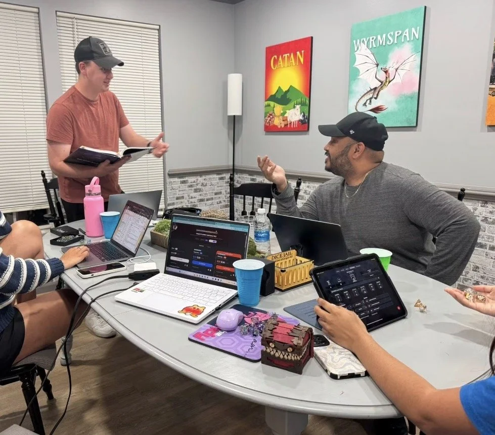 People sitting around a table with laptops and tablets, engaged in discussion in a room with game-themed artwork on the wall.
