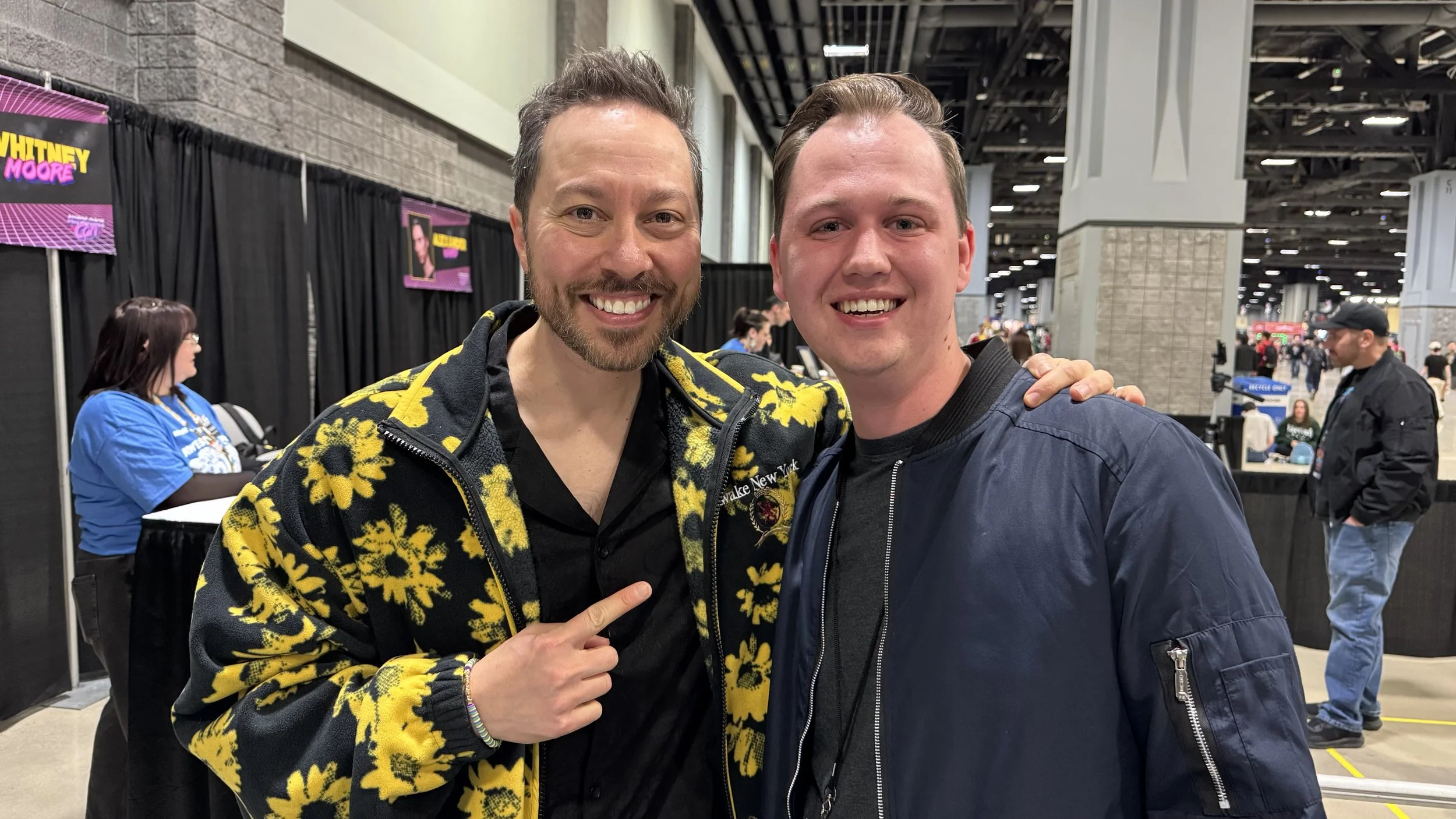 Two smiling men at an indoor event, one wearing a yellow floral jacket and the other in a black jacket, with a background of event booths and people.