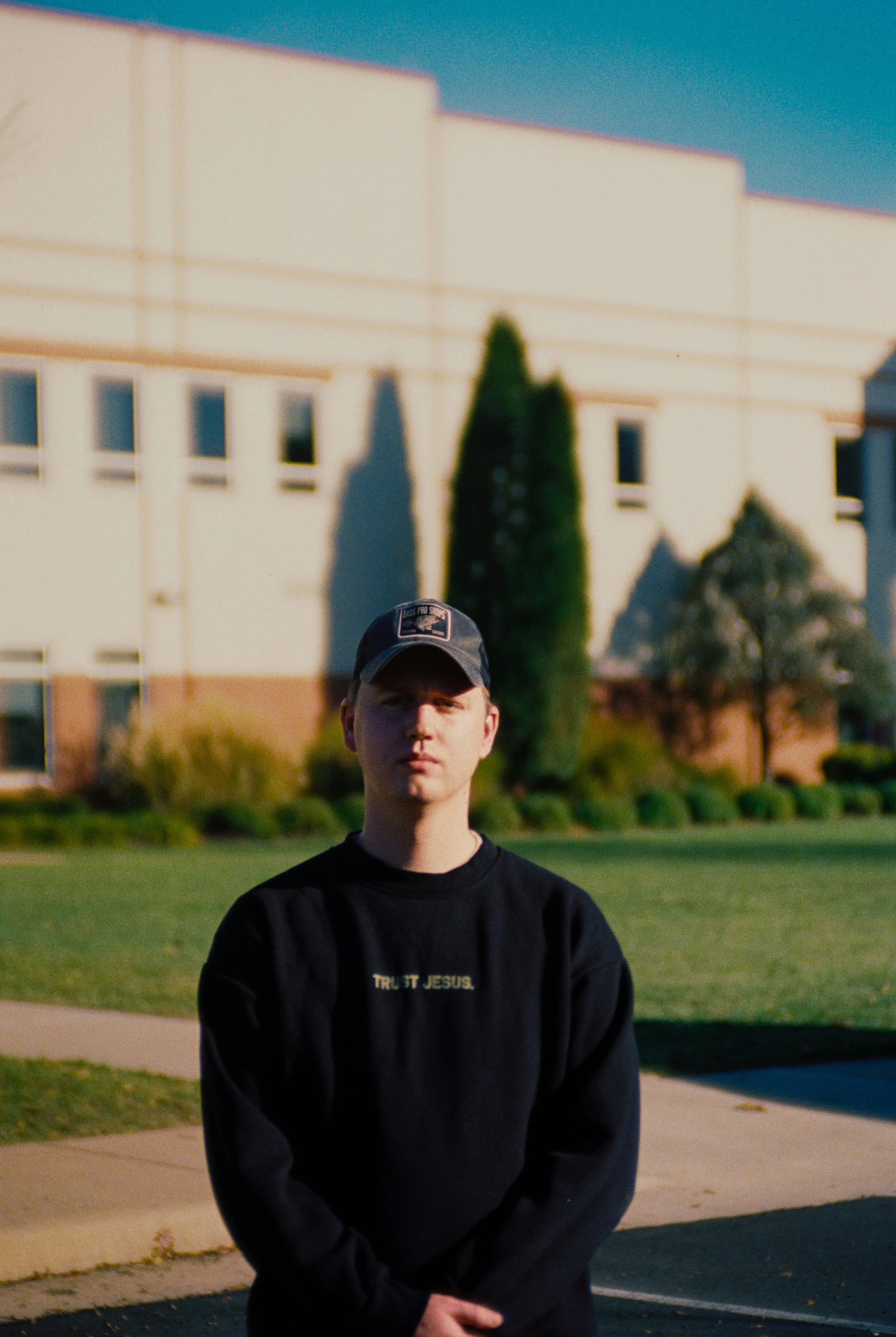 A young man wearing a black sweatshirt and a baseball cap standing outdoors in front of a building with small windows and a tall, narrow tree, during sunset.