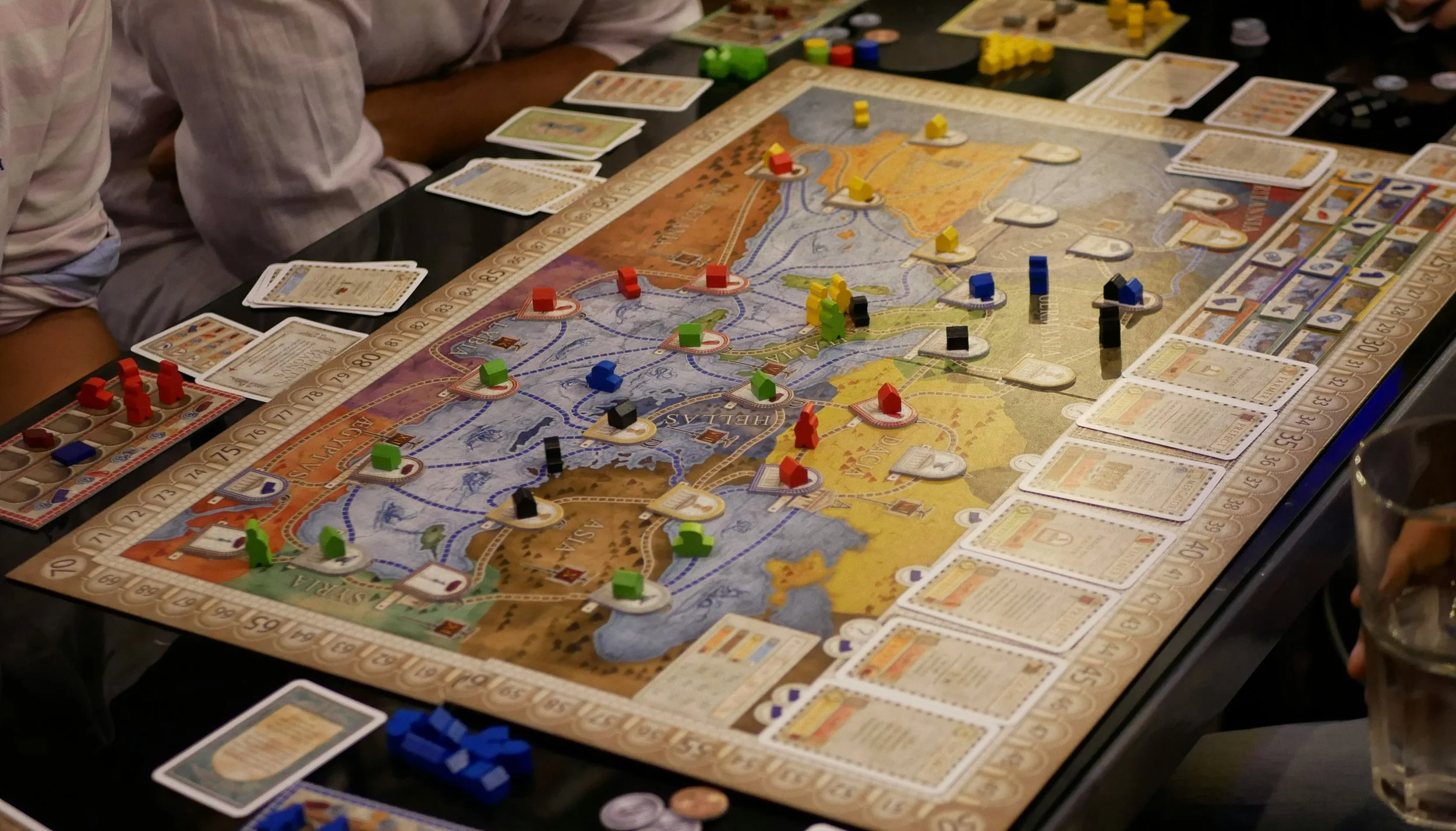 A group of people playing the board game 'Ticket to Ride' around a table. The game board features a map, various colored train pieces, and cards laid out.