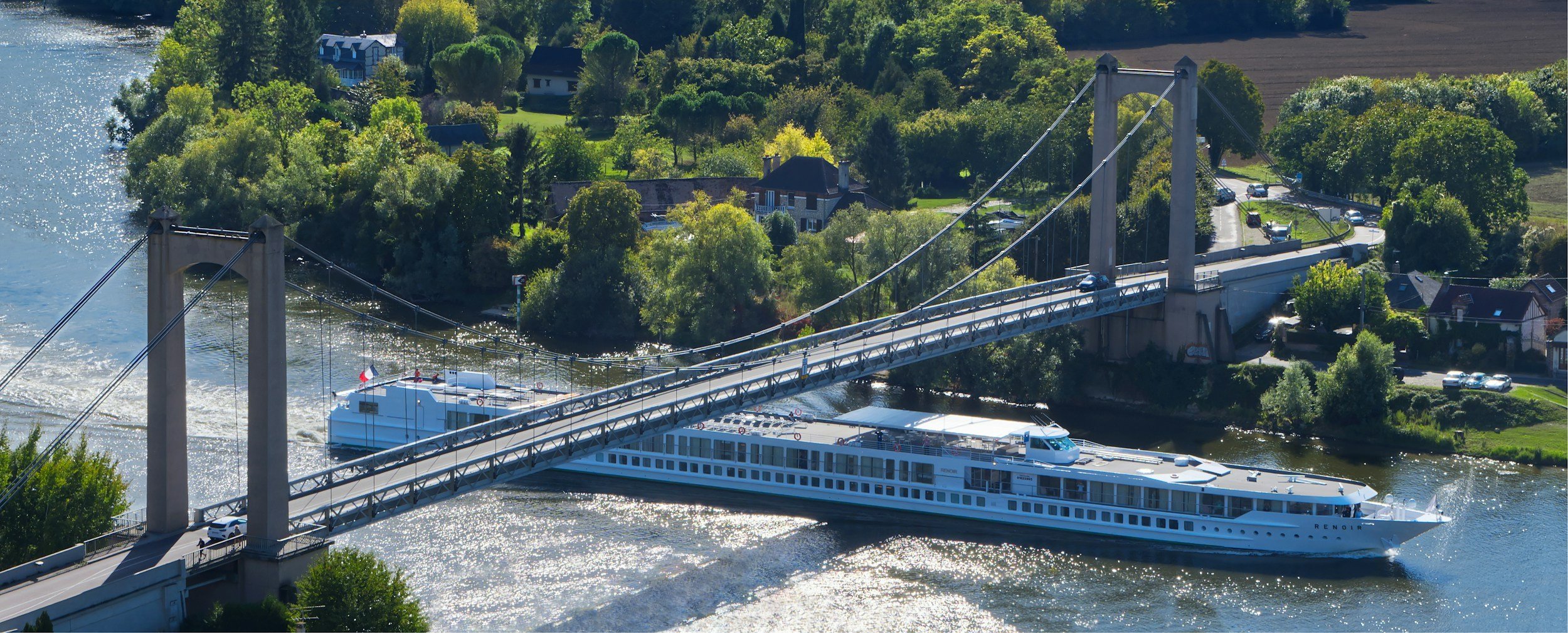 Aerial view of a suspension bridge crossing a river, with a large white cruise boat underneath. There are green trees, houses, and a parking lot along the riverbank.