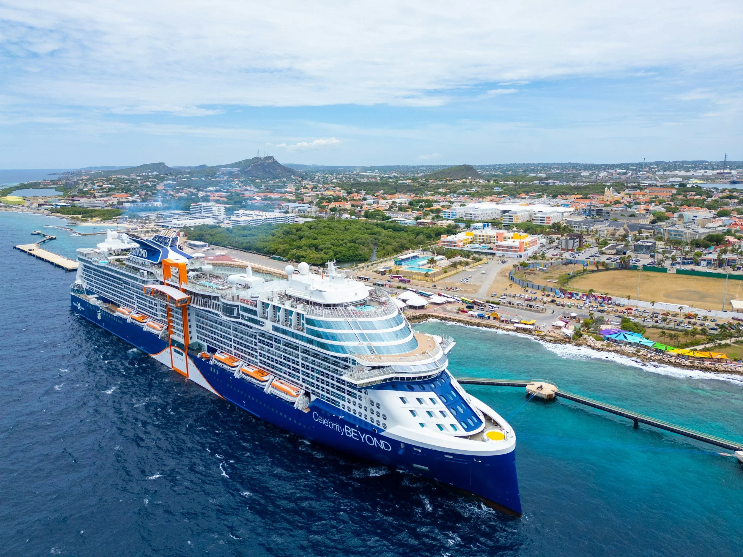 A large cruise ship docked at a port near a city with a coastline, green parks, low-rise buildings, and hills in the background under a partly cloudy sky.