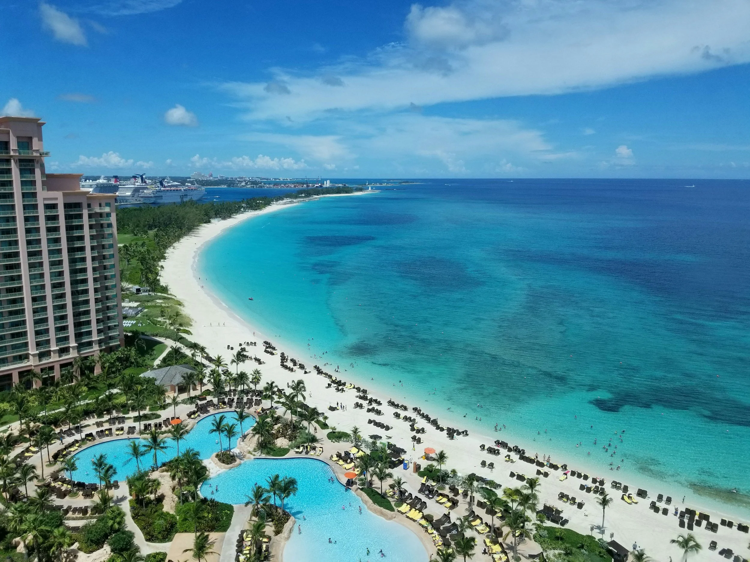 Aerial view of a beach resort with a large hotel, swimming pools, lounge chairs, palm trees, and turquoise ocean water under a partly cloudy sky.