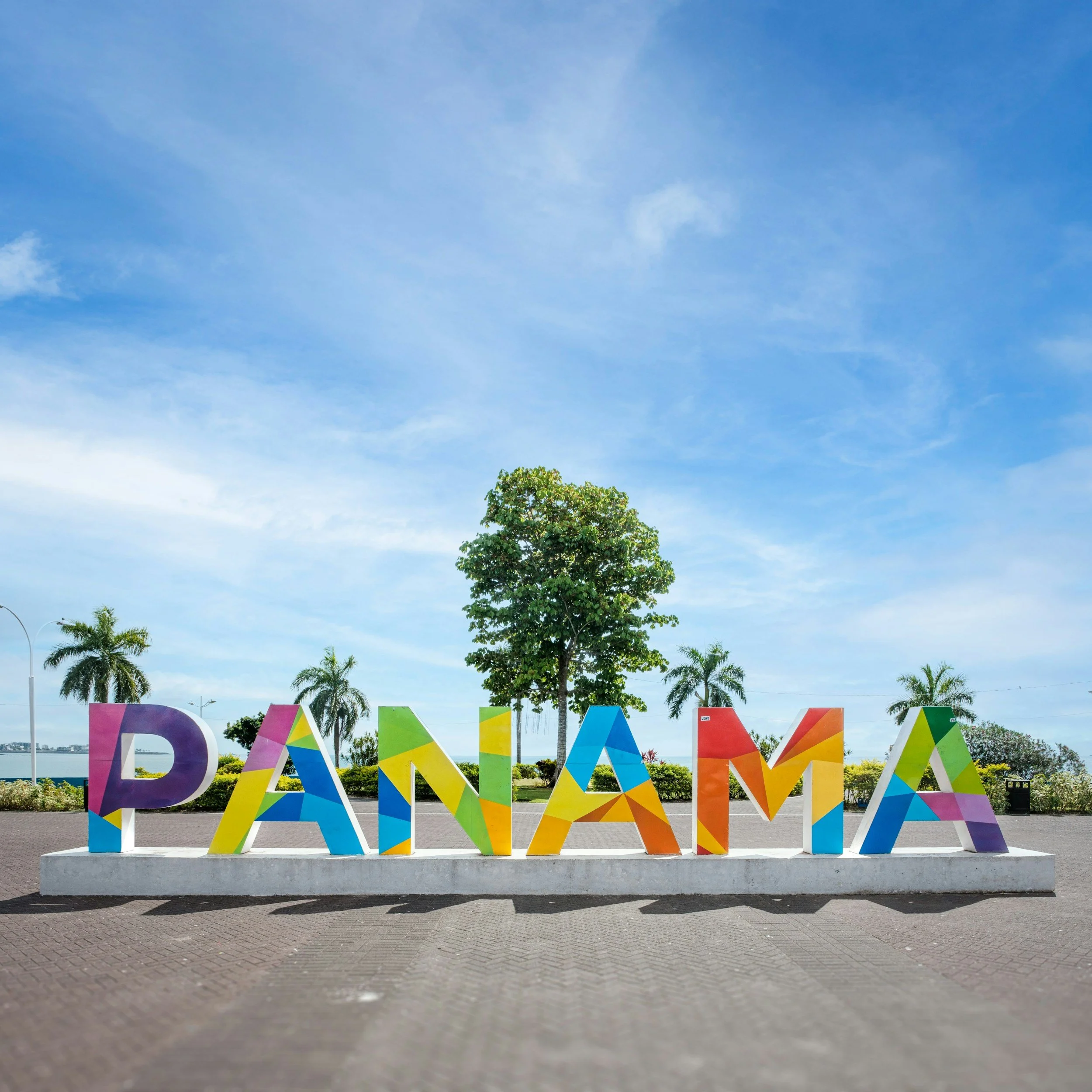 Colorful decorative letters spelling the word 'PUERTO' in an outdoor setting with a tree, some palms, and a blue sky with clouds in the background.