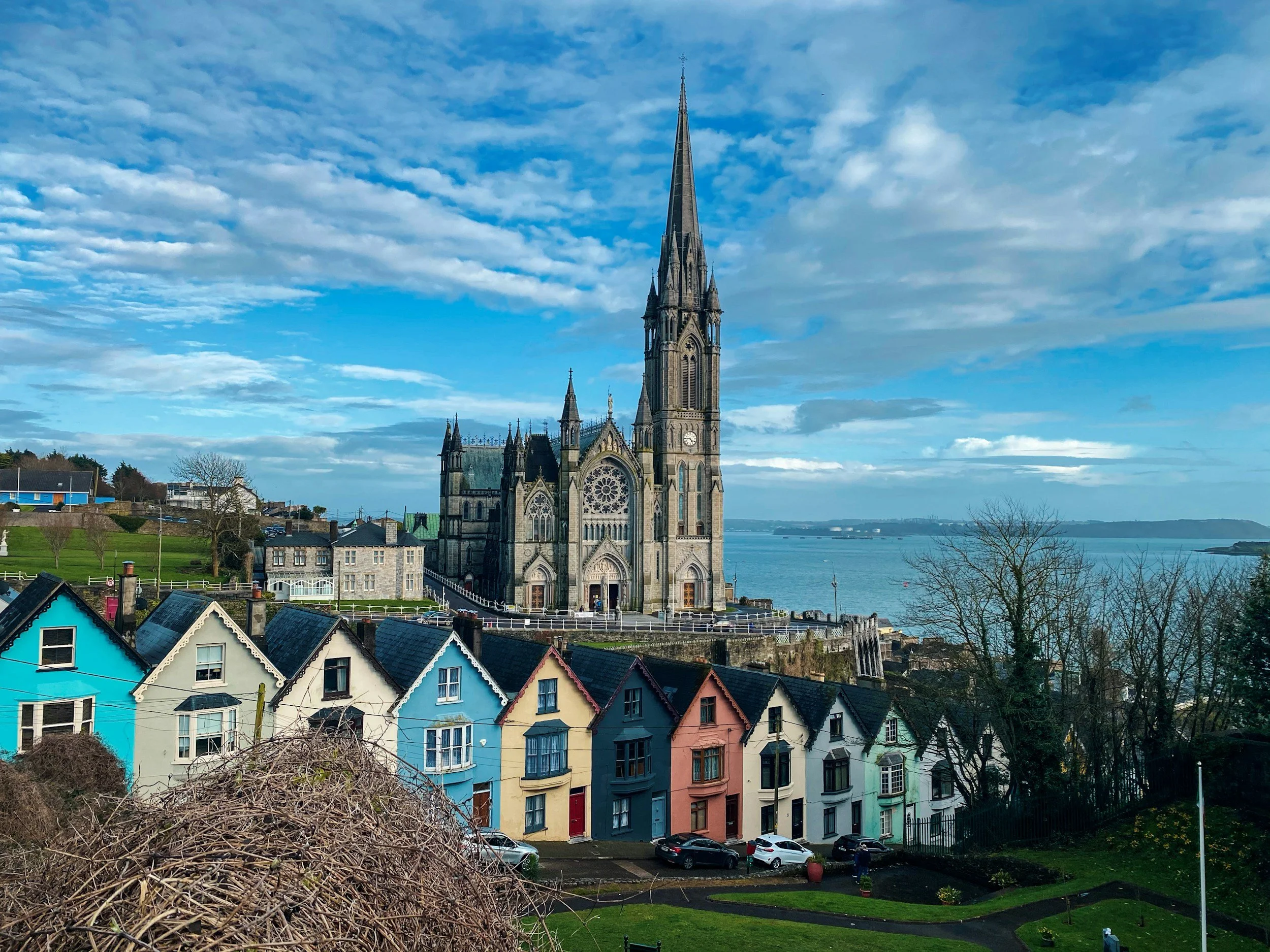 Colorful row of houses in front of a historic church with a tall spire, overlooking water with hills in the background, under cloudy sky.