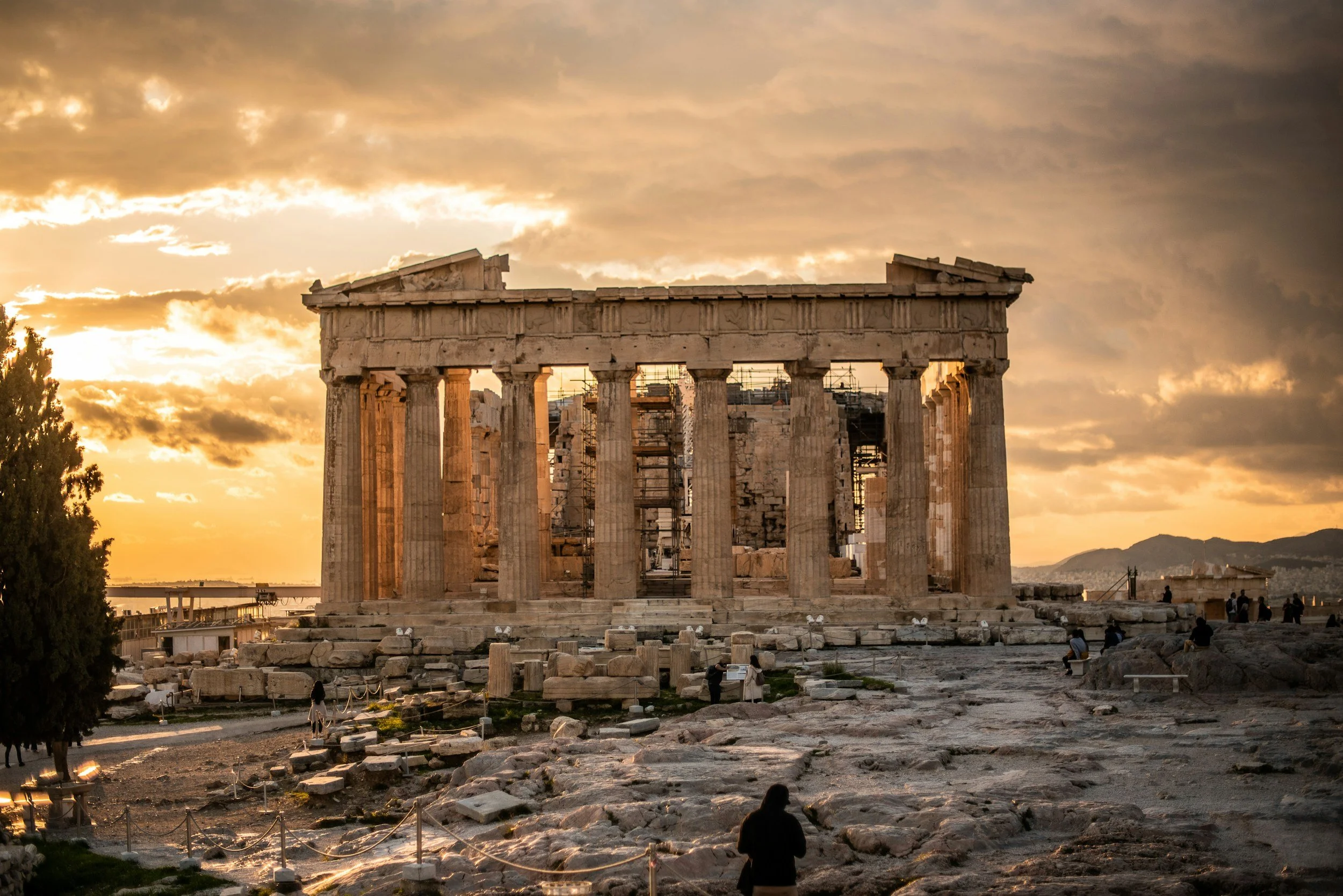 Ancient Greek Parthenon temple ruins during sunset with scattered stone blocks and a cloudy sky.