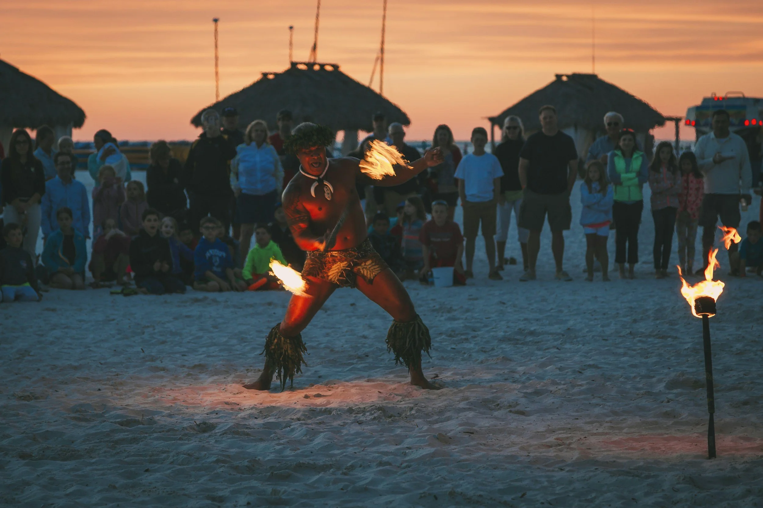 A person performing a fire dance on a sandy beach during sunset, surrounded by a crowd of onlookers.