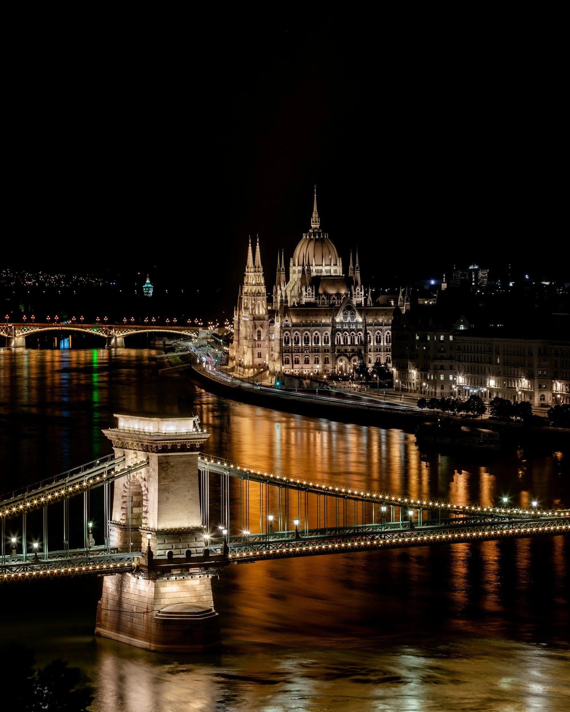 Night view of the illuminated Margaret Bridge in Budapest, Hungary, with the Parliament building and the Danube River reflected in the water.