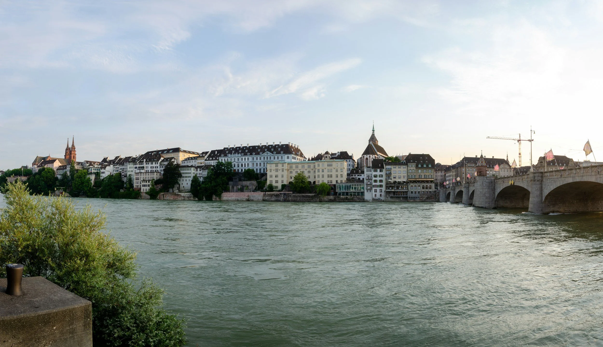 View of a city skyline across a river with historic buildings, church steeples, and a stone bridge, under a partly cloudy sky.