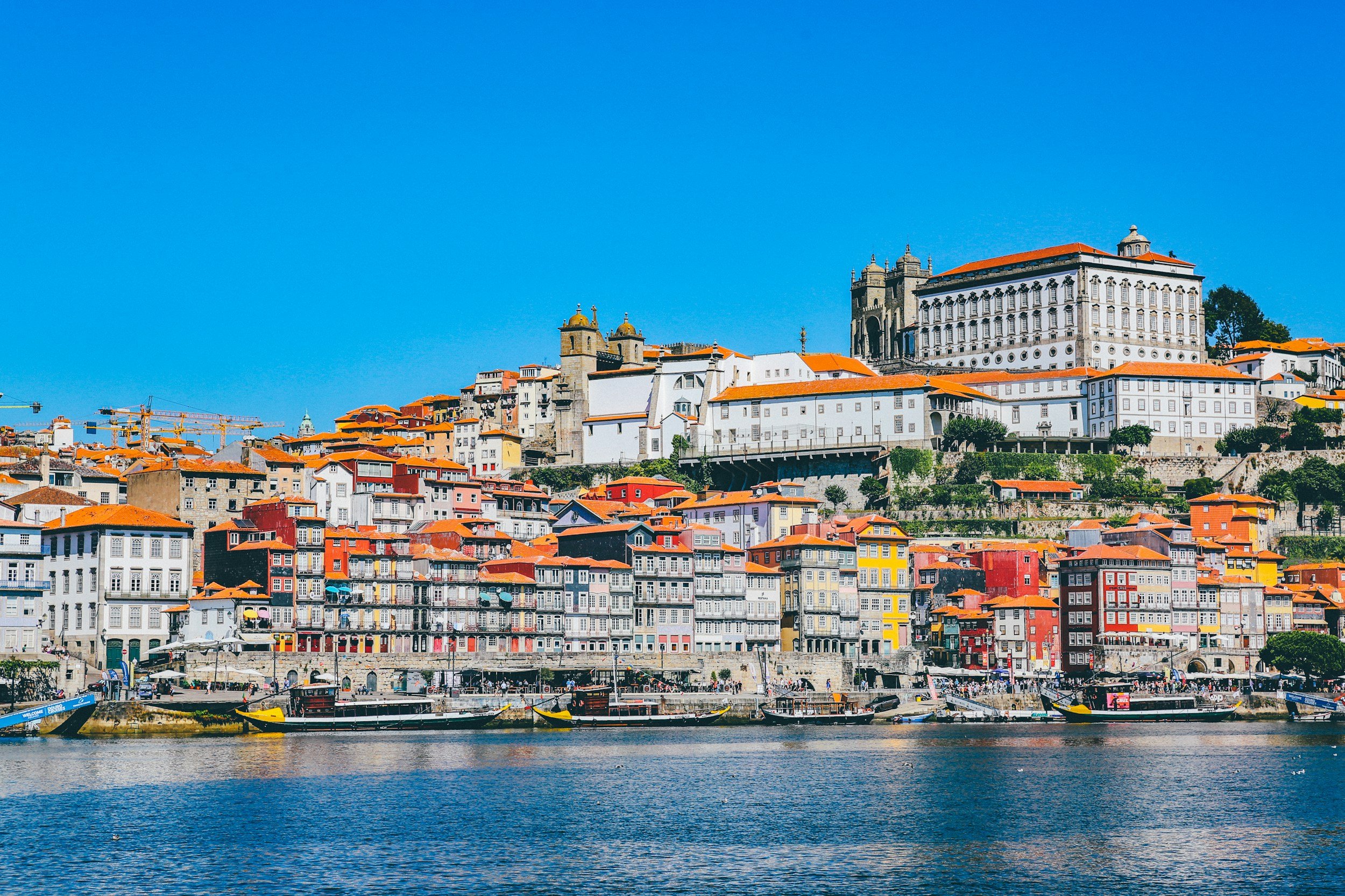 Colorful houses with orange roofs along the riverside, with historic buildings and a church on a hillside against a clear blue sky.