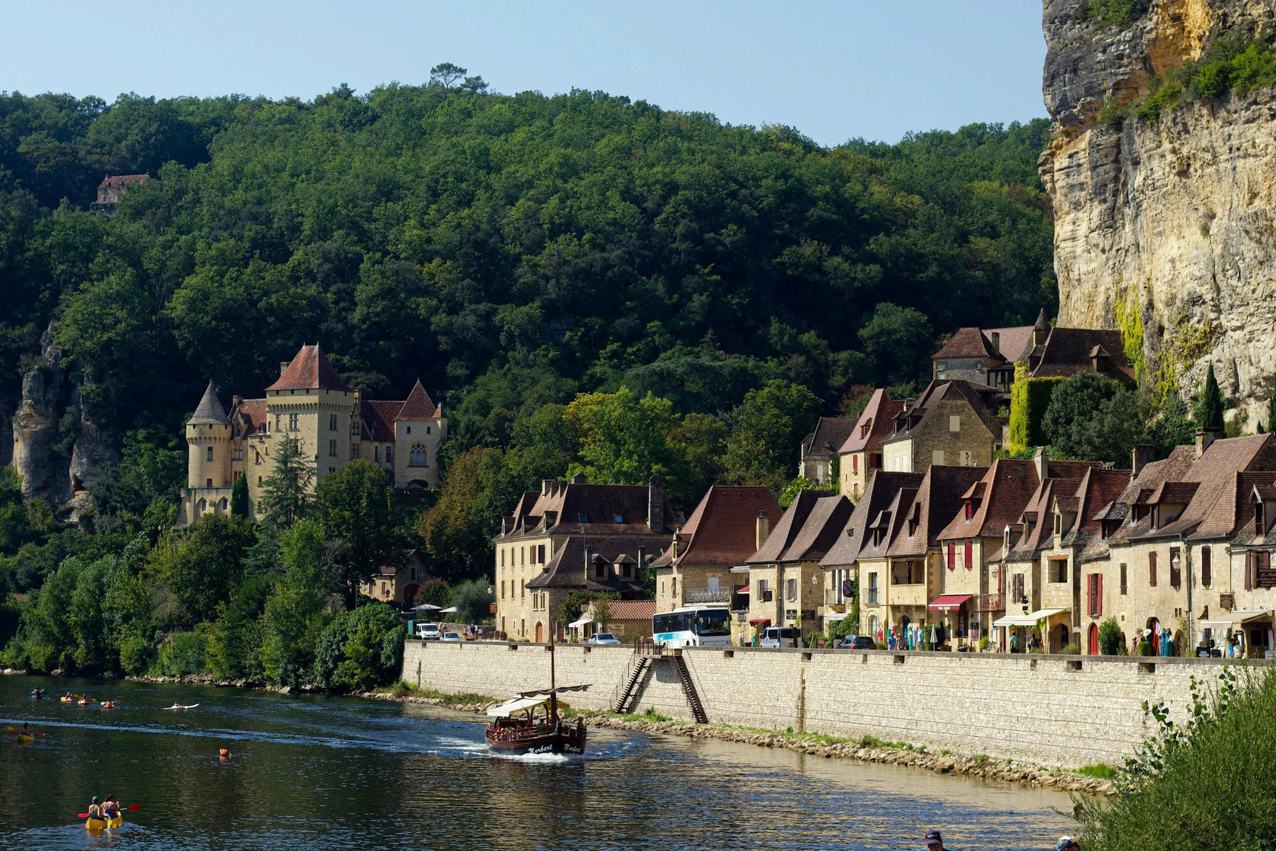 A scenic view of a riverside town with historic buildings, a castle, and a castle tower nestled among lush green hills with a rocky cliff face on the right. People are kayaking and boating on the river.