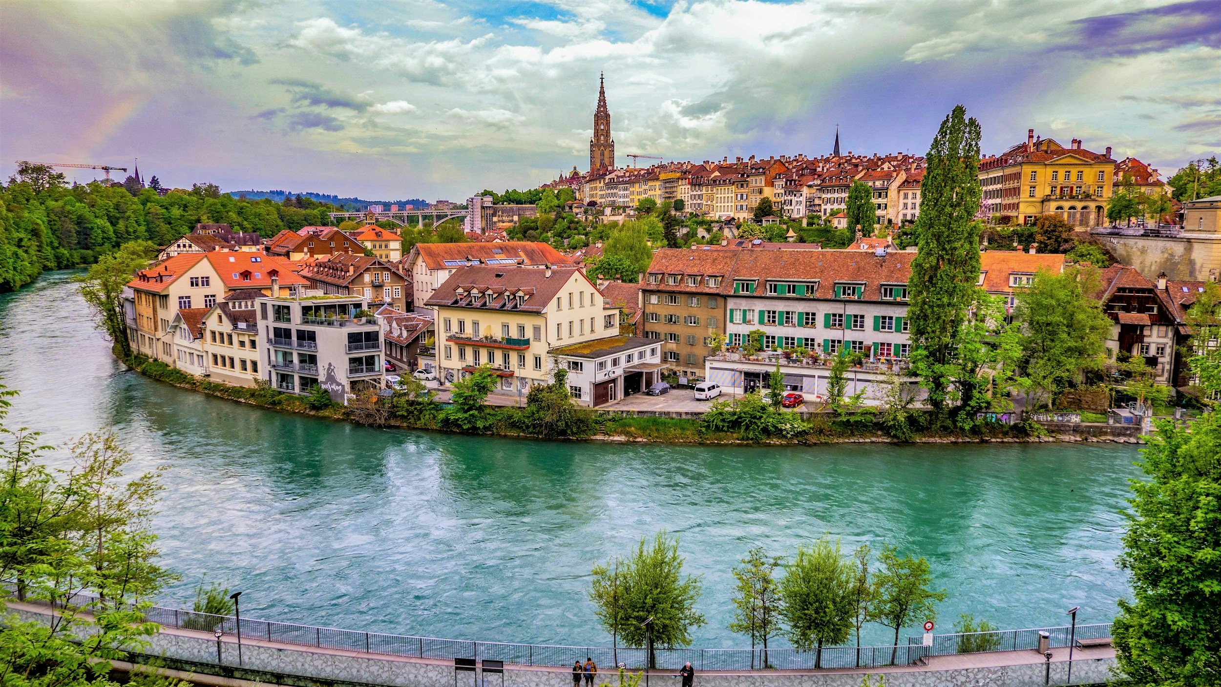 A scenic view of a river with greenish water flowing through a city with buildings and houses along its banks. In the background, there are many colorful buildings with a tall church steeple rising above them, set against a sky with clouds and a rainbow.