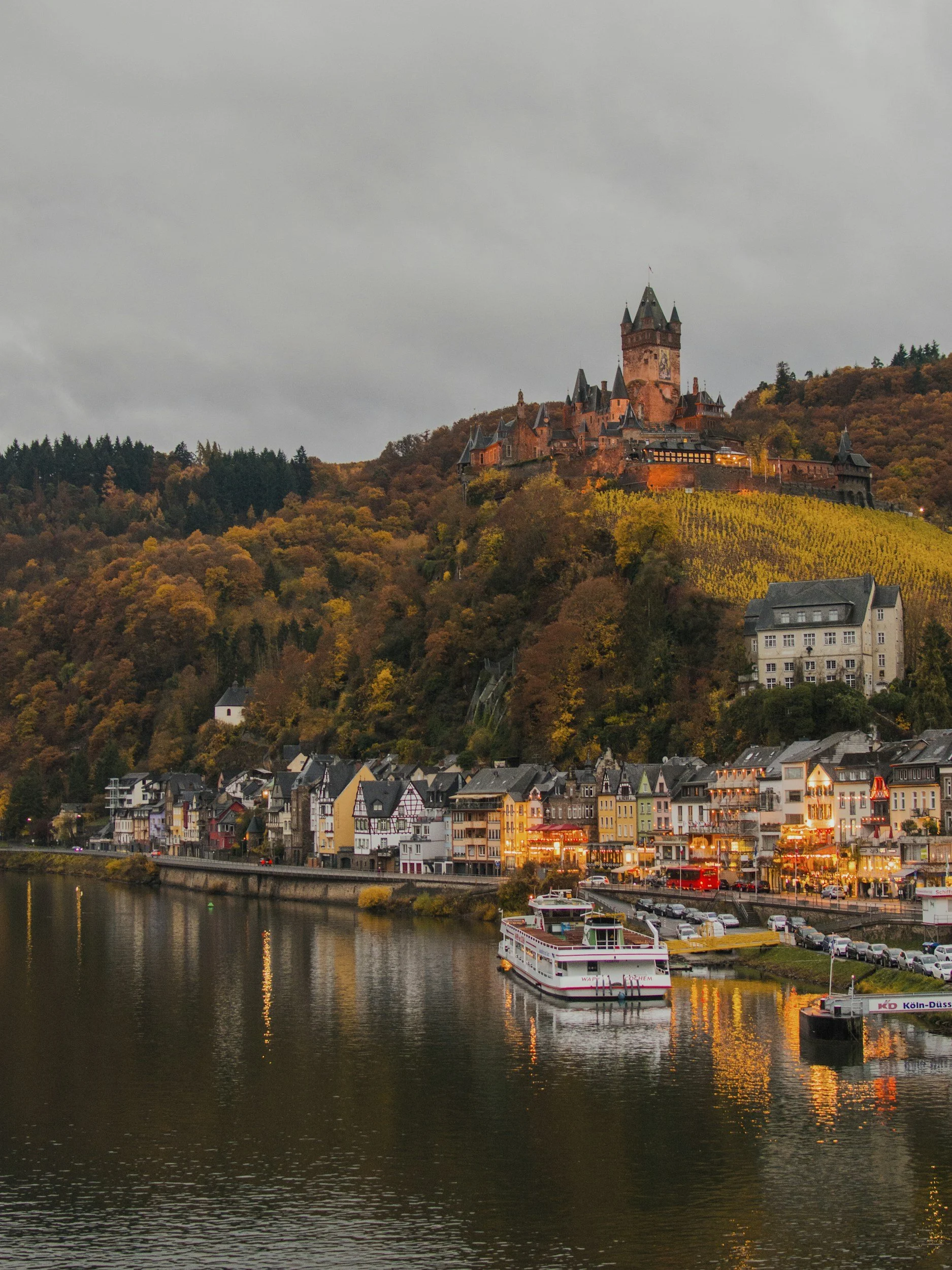 A riverside town with colorful houses lining the water, a boat in the river, and a castle on a hill in the background during dusk.