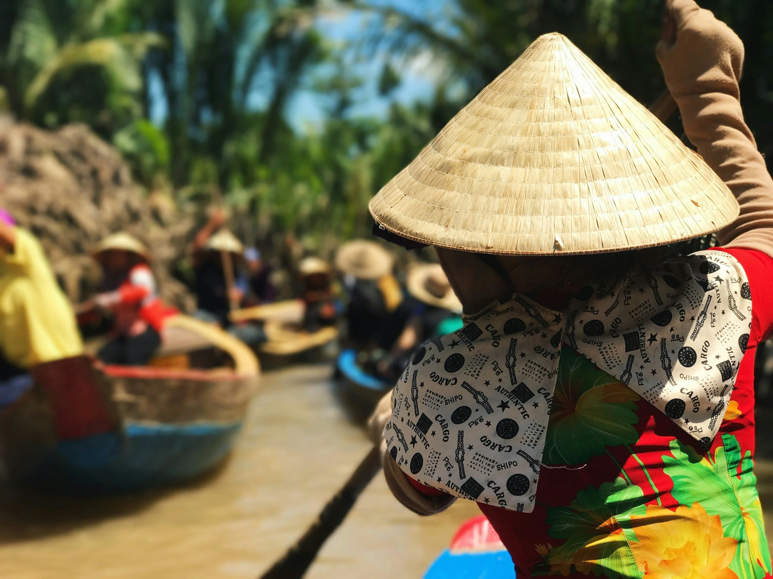 A person wearing a traditional conical hat, colorful shorts, and a patterned shirt, likely working or participating in an outdoor activity, with others in the background also wearing conical hats in a rural or agricultural setting.