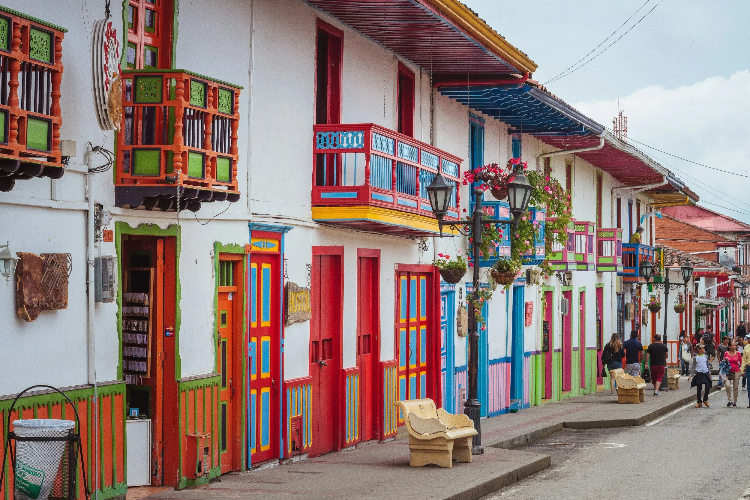 Colorful row of buildings with decorated balconies and doors, featuring bright red, green, blue, pink, and yellow accents, along with hanging flower baskets, on a busy street with pedestrians.