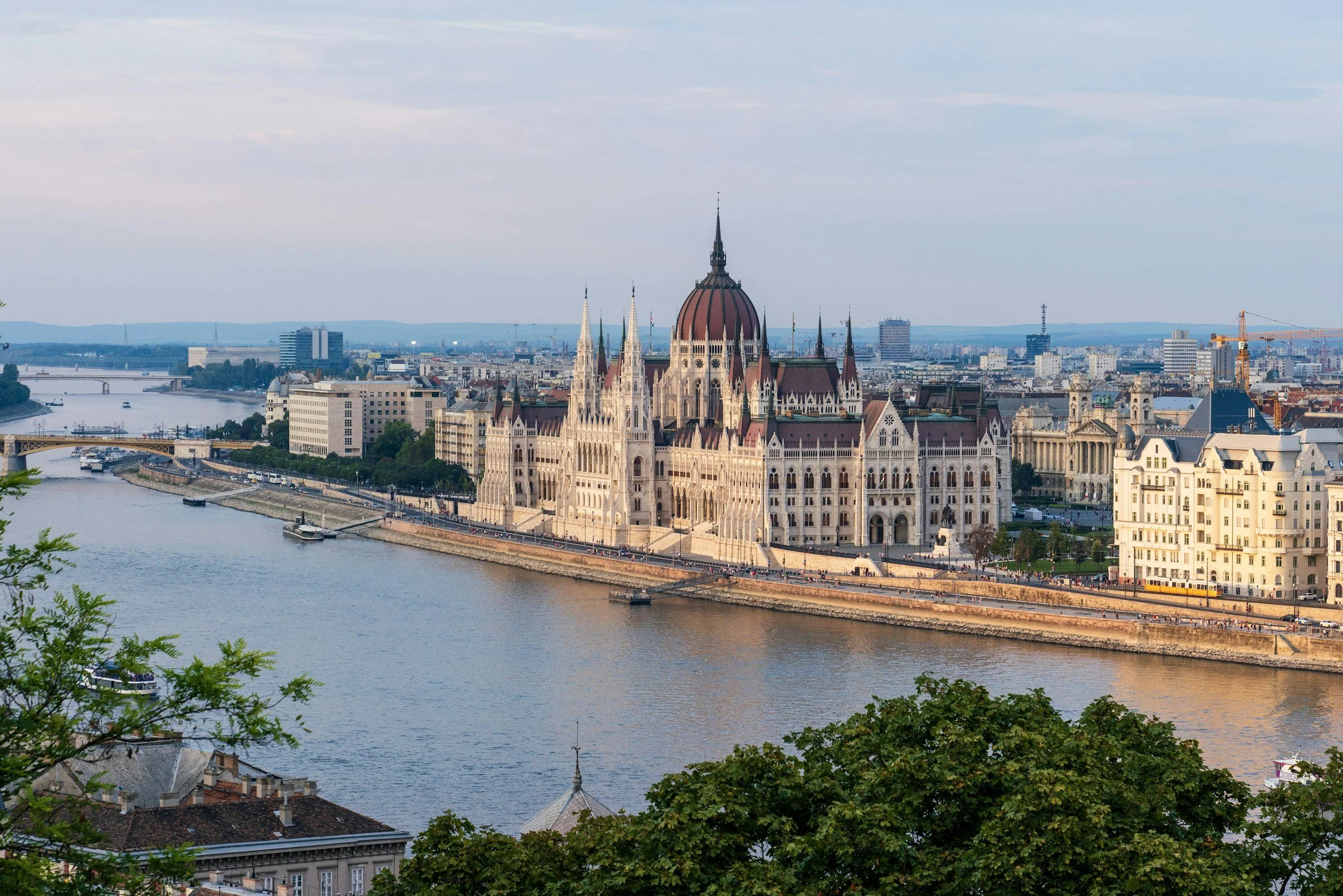 A panoramic view of Budapest skyline featuring the Parliament Building along the Danube River, with bridges and boats on the river, and a cityscape with modern and historic buildings in the background.