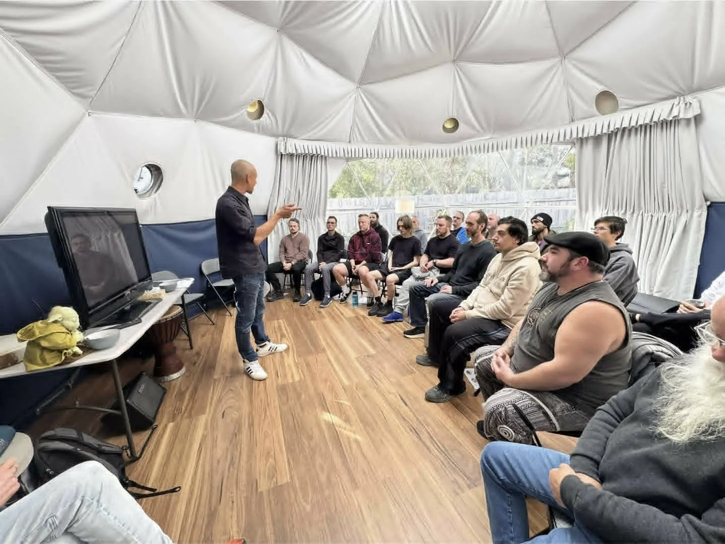 Indoor presentation with a group of people seated in a semi-circle, audience listening to a speaker standing in front of a large monitor and TV, inside a tent-like structure with white fabric walls and ceiling, wooden floor, and windows showing trees