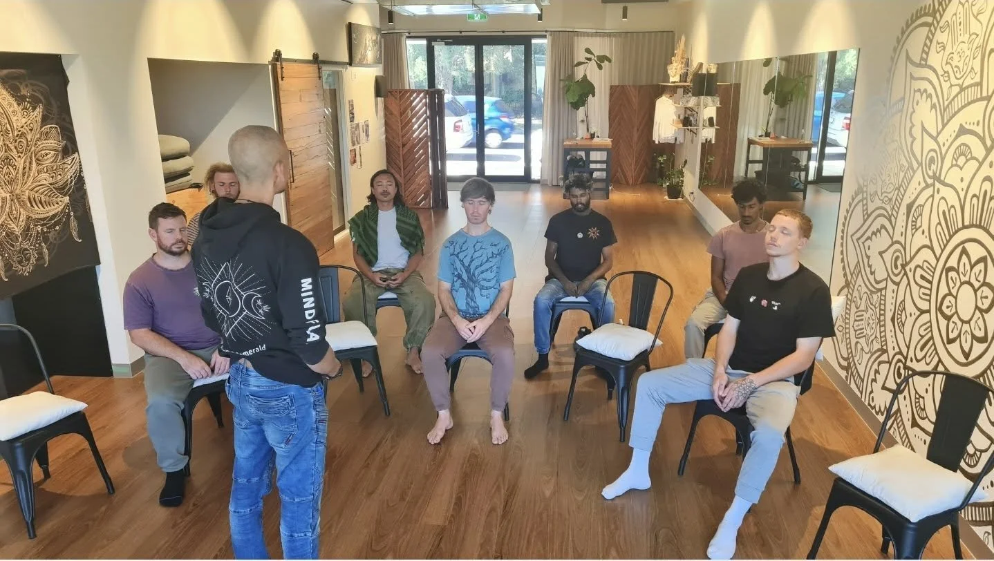A group meditation session in a bright, spacious room with wooden flooring and minimal decor. Seven people sit on black chairs arranged in a semi-circle, with one person standing and leading the session. Participants are relaxed, with some sitting ba