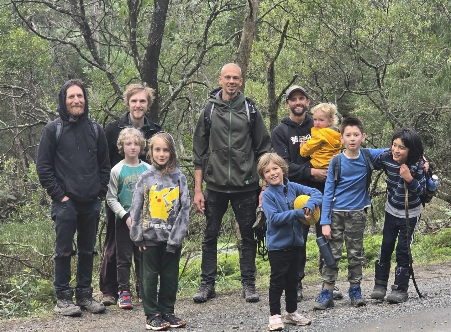 A group of ten people, including children and adults, standing outdoors in a wooded area on a forest trail during daytime. They are dressed in casual outdoor clothing and carrying backpacks and sports equipment.