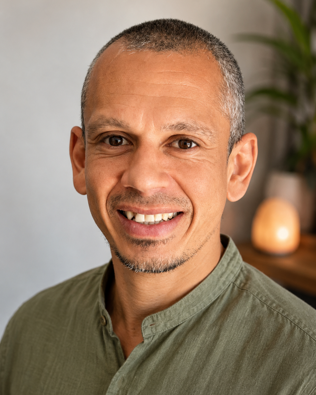 Close-up portrait of smiling middle-aged man with short gray hair, facial hair, and wearing a green shirt, indoors with a blurred background and a salt lamp.