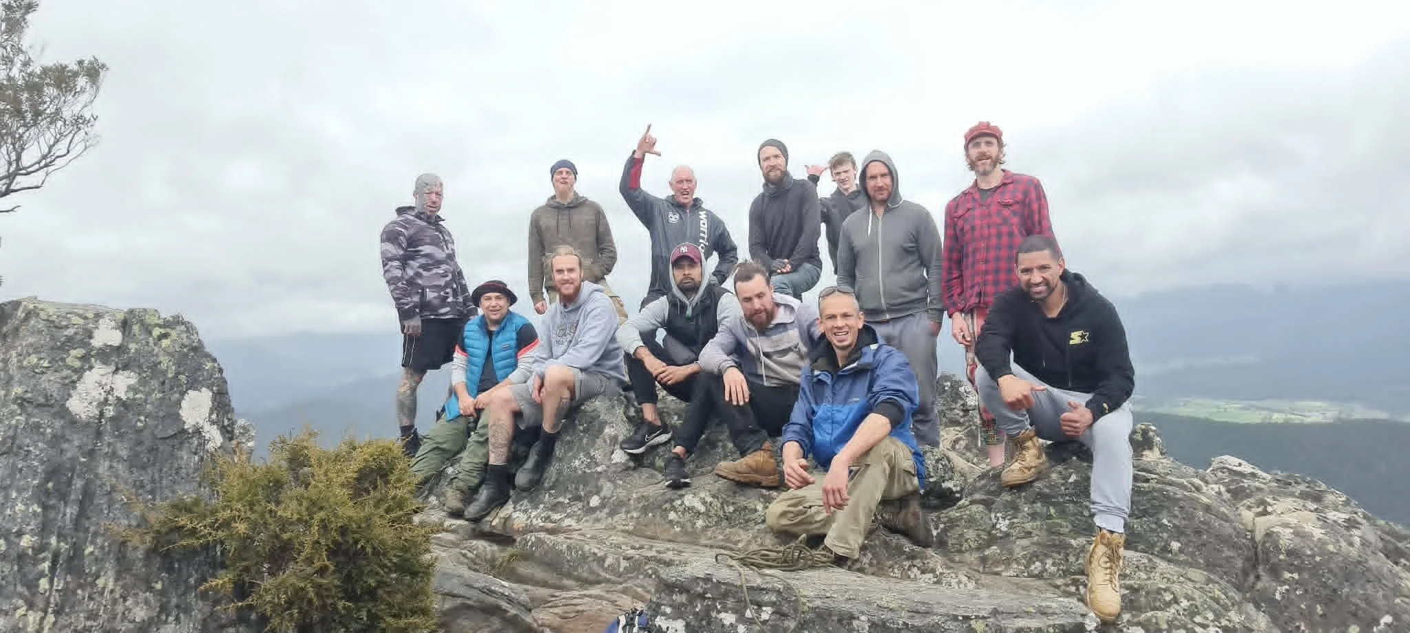 A group of friends on a mountain top, posing for a photo with a scenic valley and cloudy sky in the background. They are dressed in outdoor hiking gear.